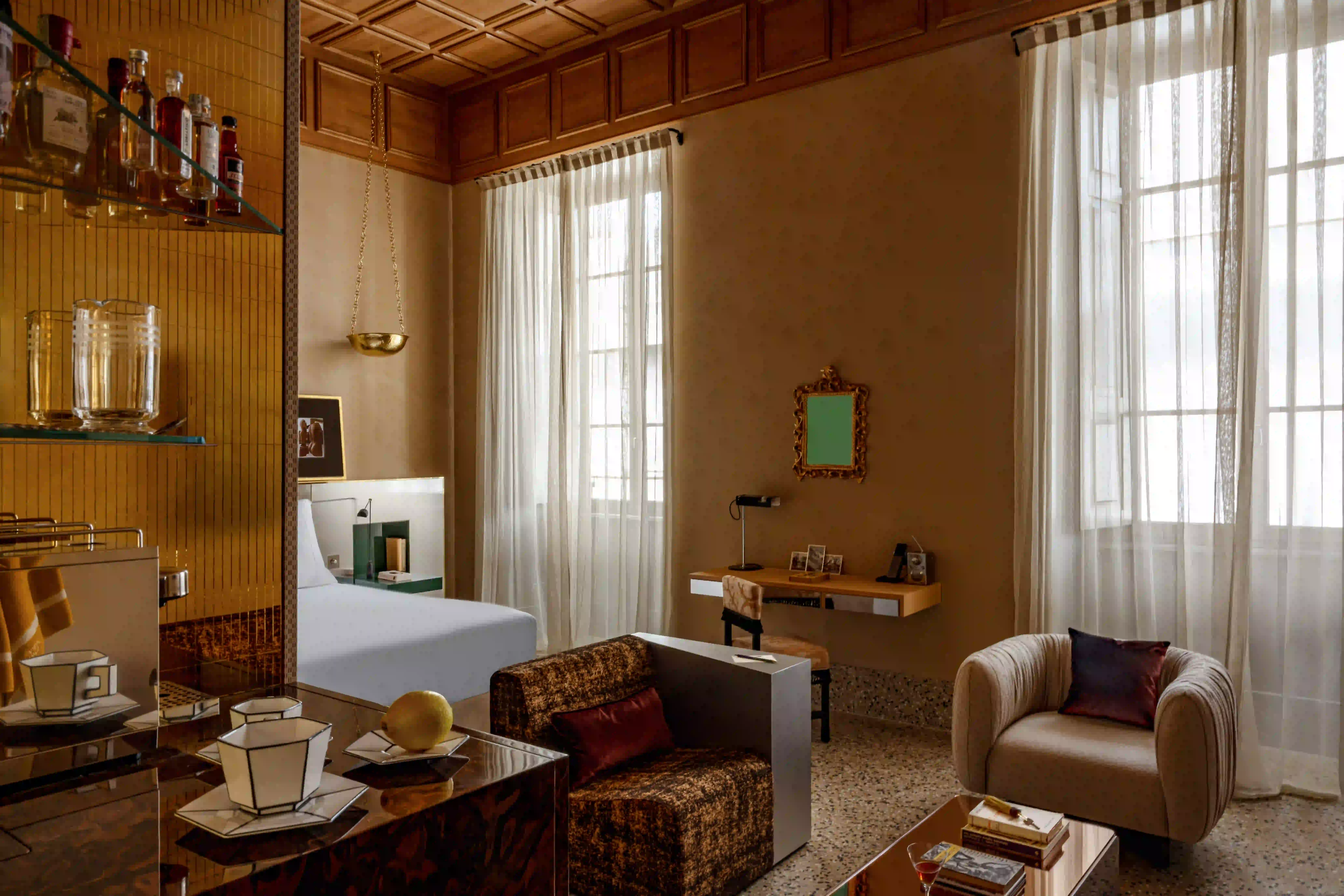 Quarto aconchegante com uma cama, duas poltronas, uma mesa, cortinas transparentes e uma prateleira com garrafas e copos.