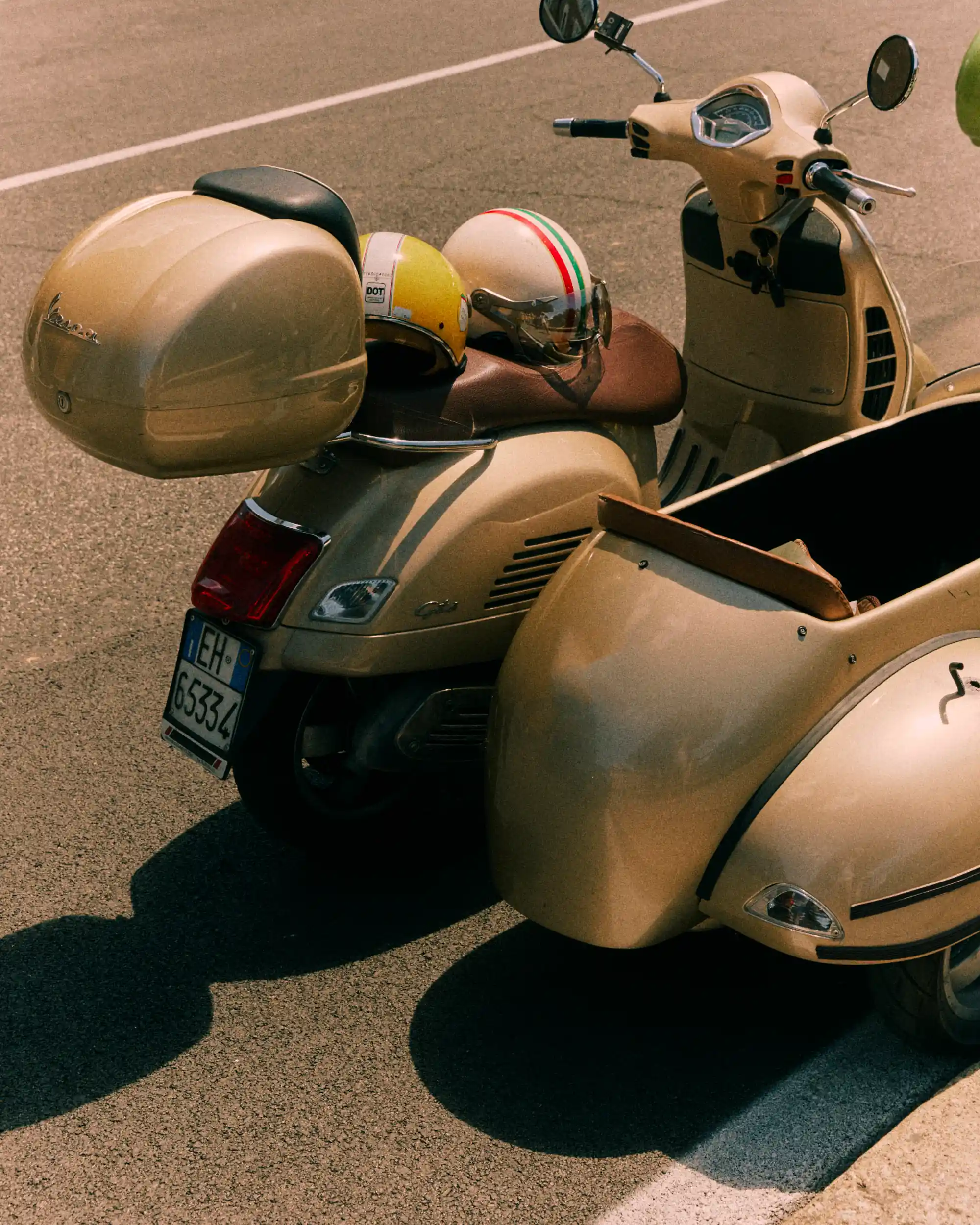 Patinete Vespa beige con sidecar y dos cascos estacionado en una carretera asfaltada.