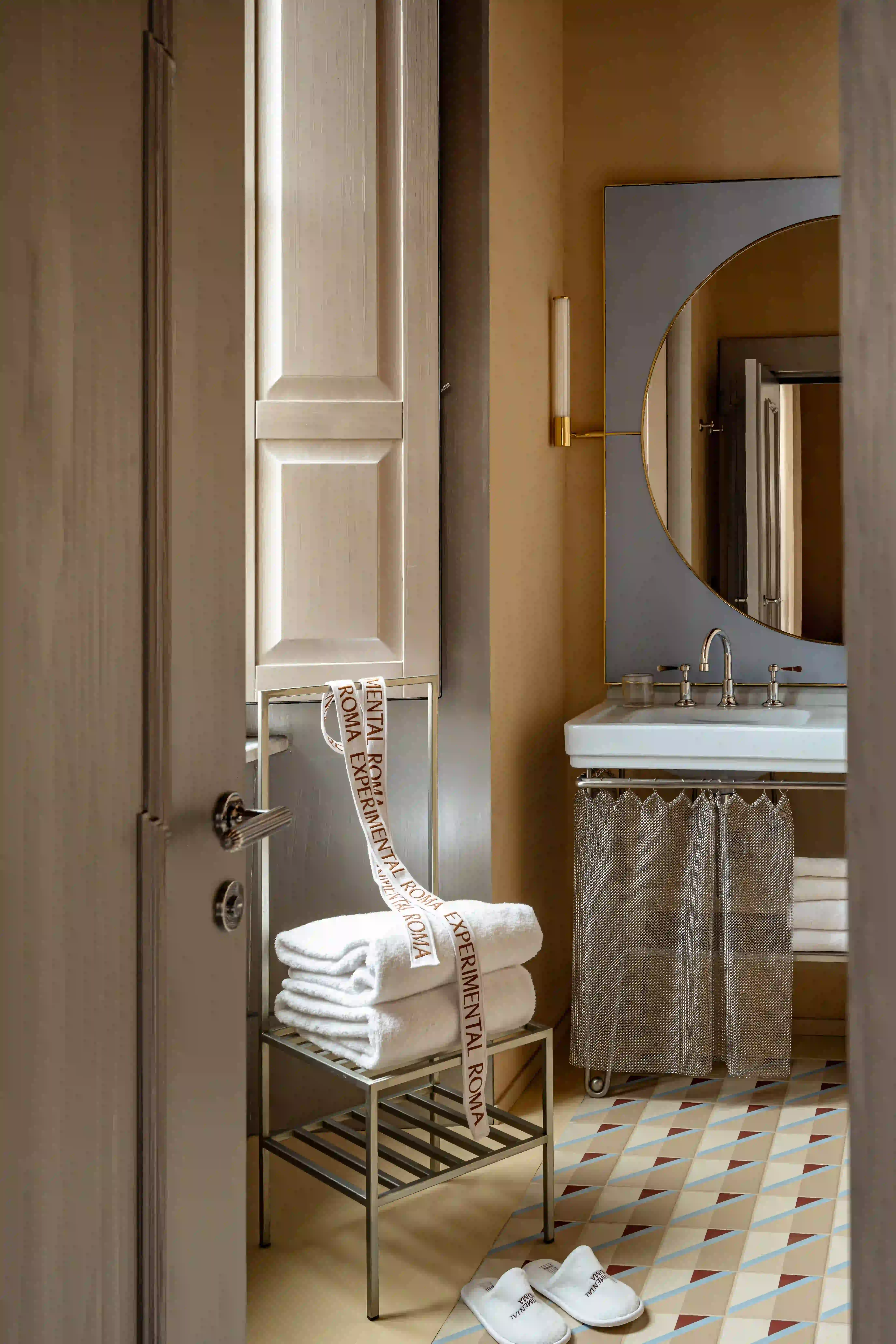 Serviettes blanches empilées sur une chaise en métal avec ruban Experimental Roma dans une salle de bain aux tons chauds.