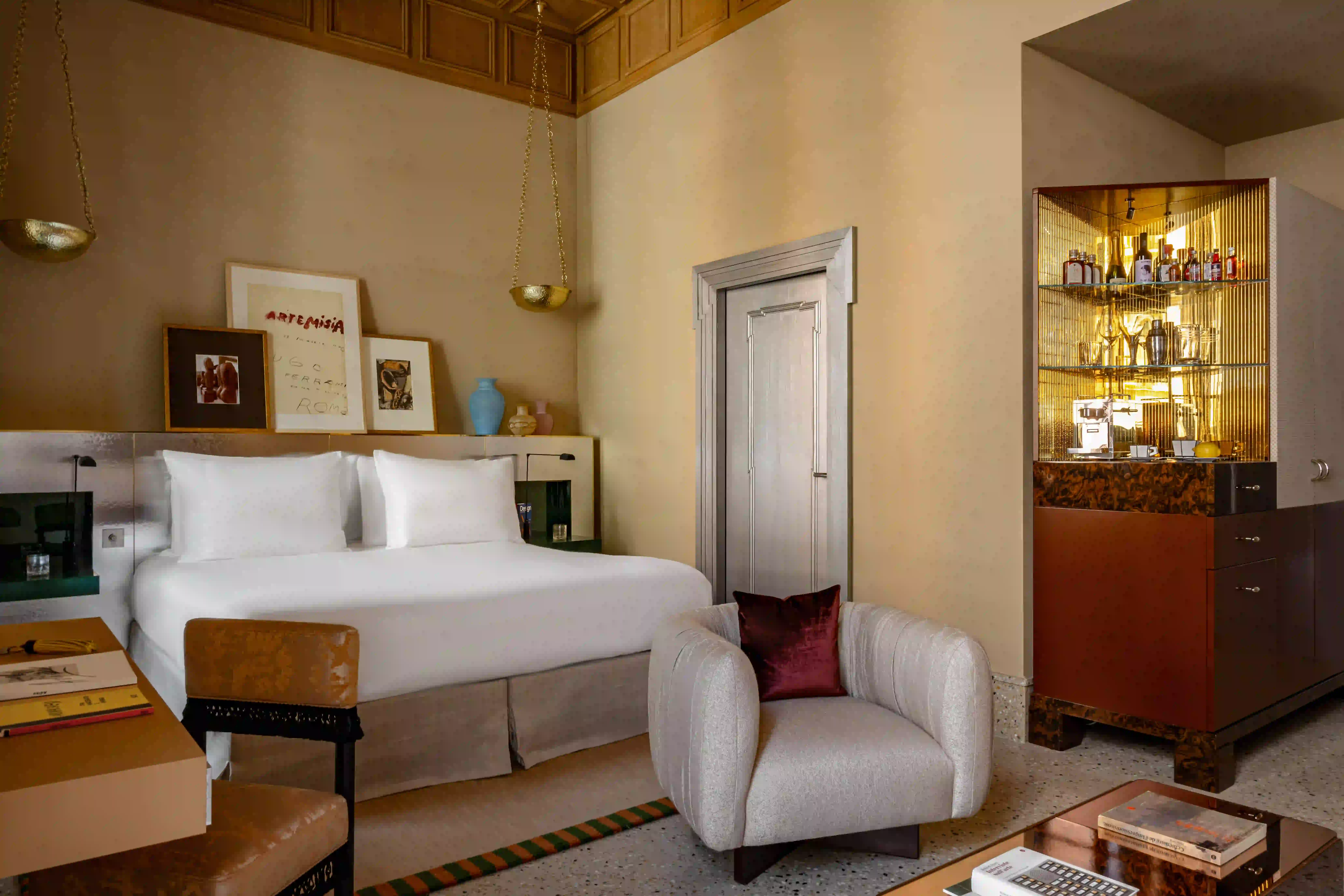 Chambre d'hôtel confortable avec un grand lit blanc, un fauteuil avec oreiller rouge et un minibar avec bouteilles.