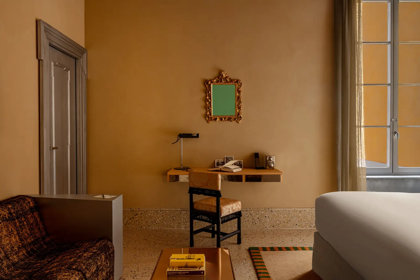 Camera minimalista con pareti beige, una piccola scrivania con sedia, specchio decorato e grande finestra con tende trasparenti.