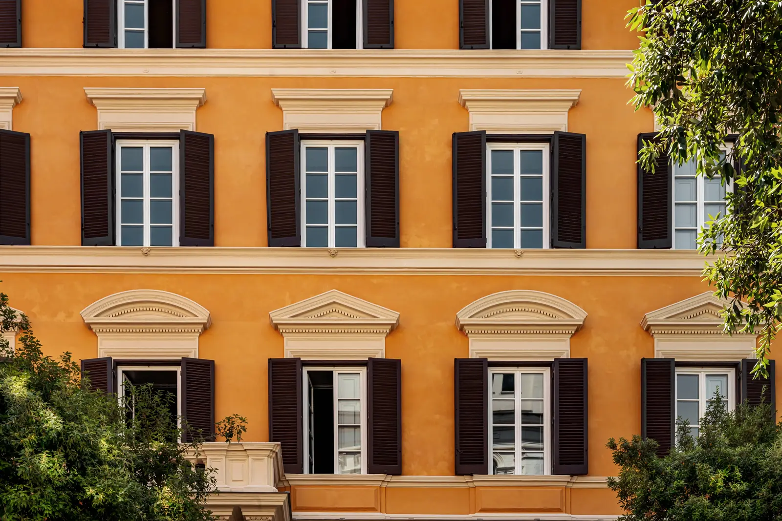 Facciata arancione dell'edificio con finestre simmetriche con cornice bianca e persiane scure parzialmente aperte, alberi intorno.
