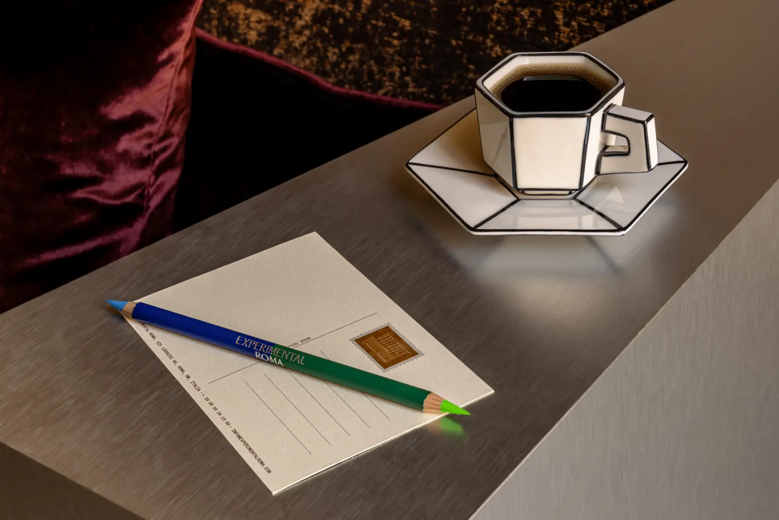 Tazza ottagonale di caffè nero su piattino accanto a cartolina e matita bicolore su un tavolo grigio.