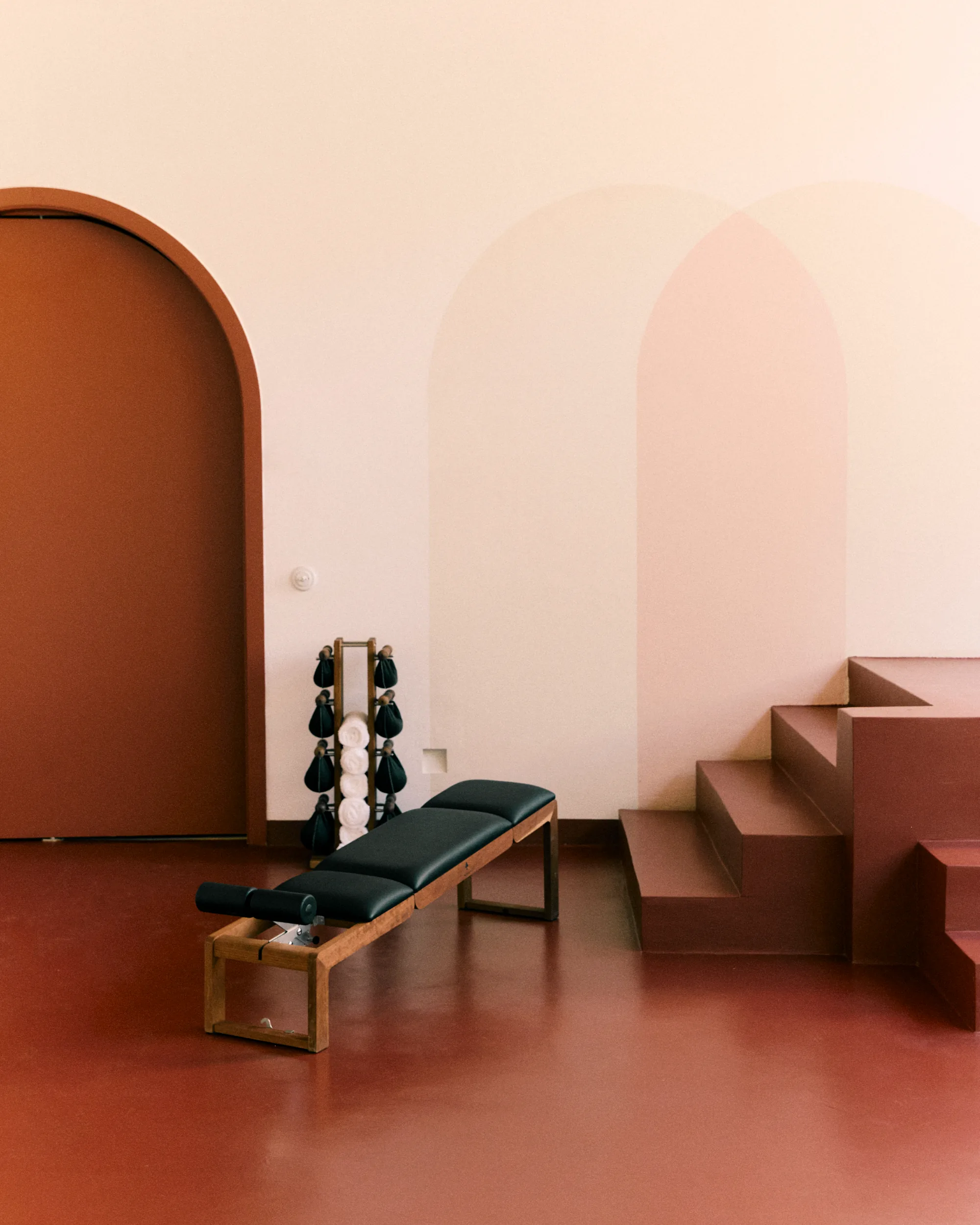 Sala de ginástica minimalista com piso marrom, porta em arco, banco preto e pesos em um rack.