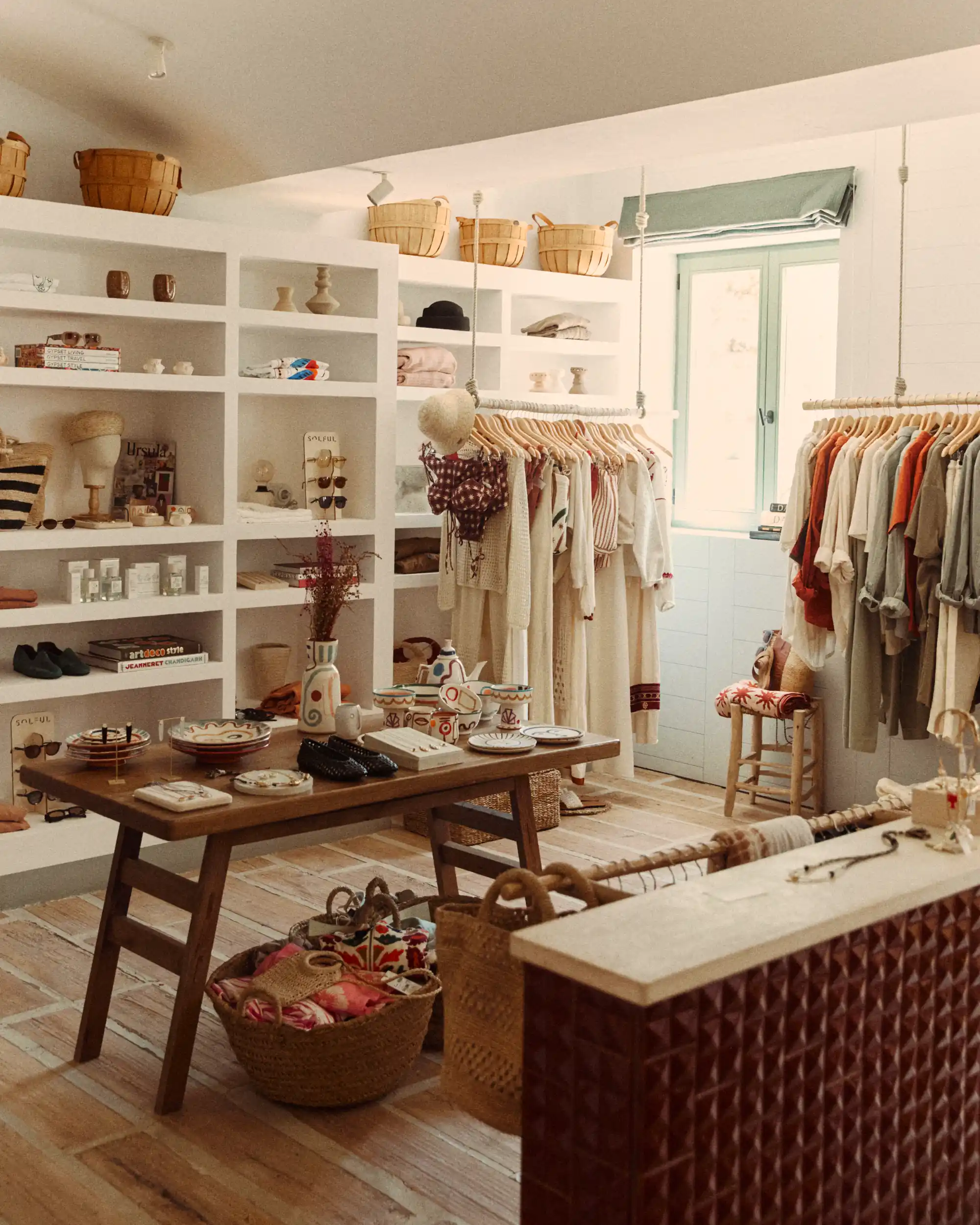 Boutique com roupas penduradas, mesa de madeira com cerâmica, cestos e prateleiras com decoração e roupas.