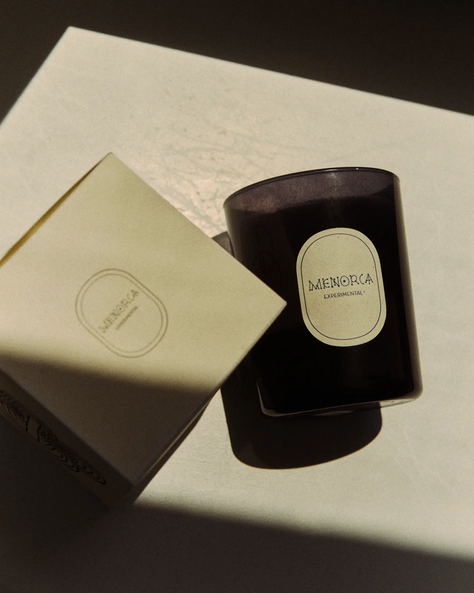 Um recipiente de vela escura com o rótulo Menorca Experimental ao lado de sua caixa de cor creme com etiqueta correspondente.