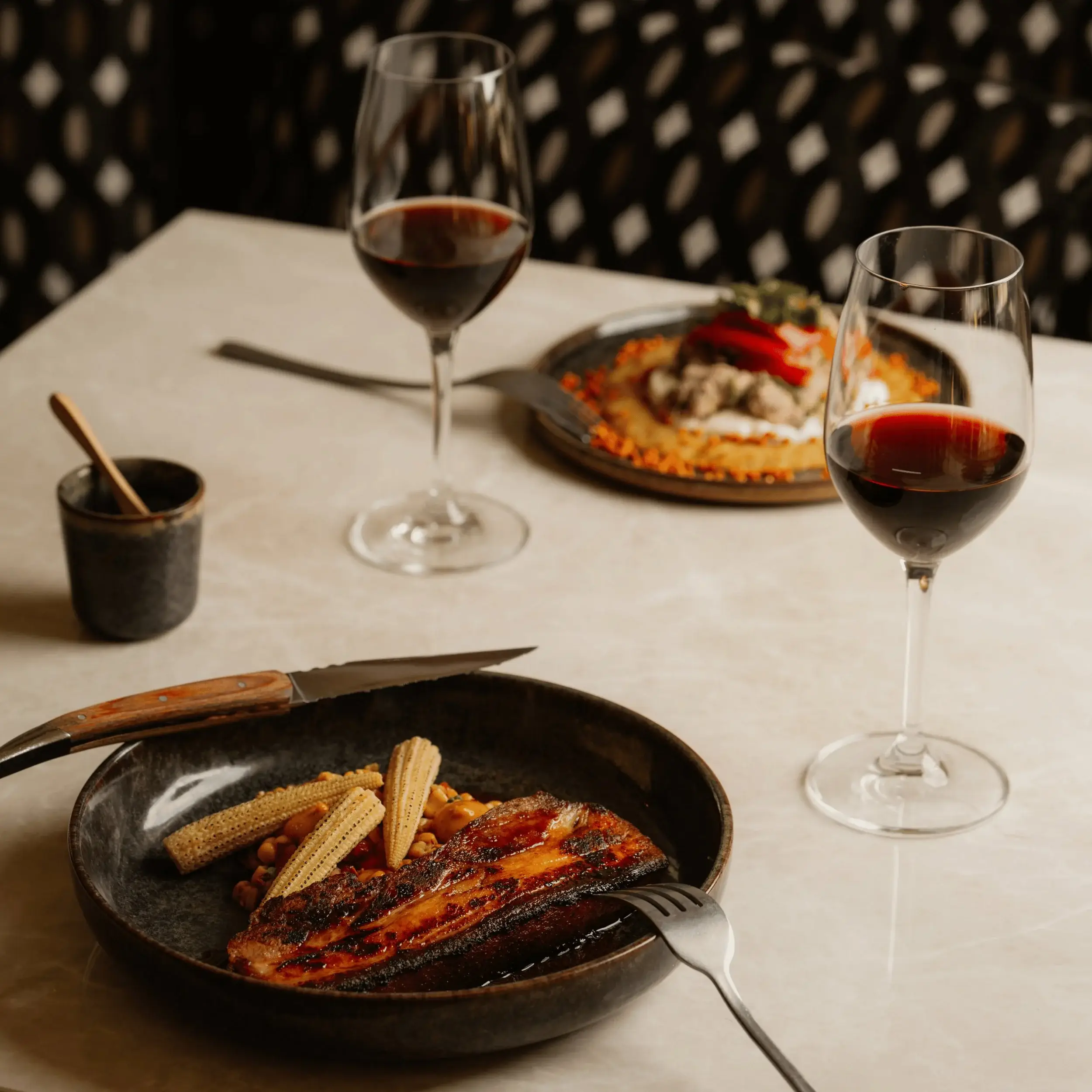 Dos platos con comida gourmet y dos copas de vino tinto sobre una mesa de restaurante de color beige.