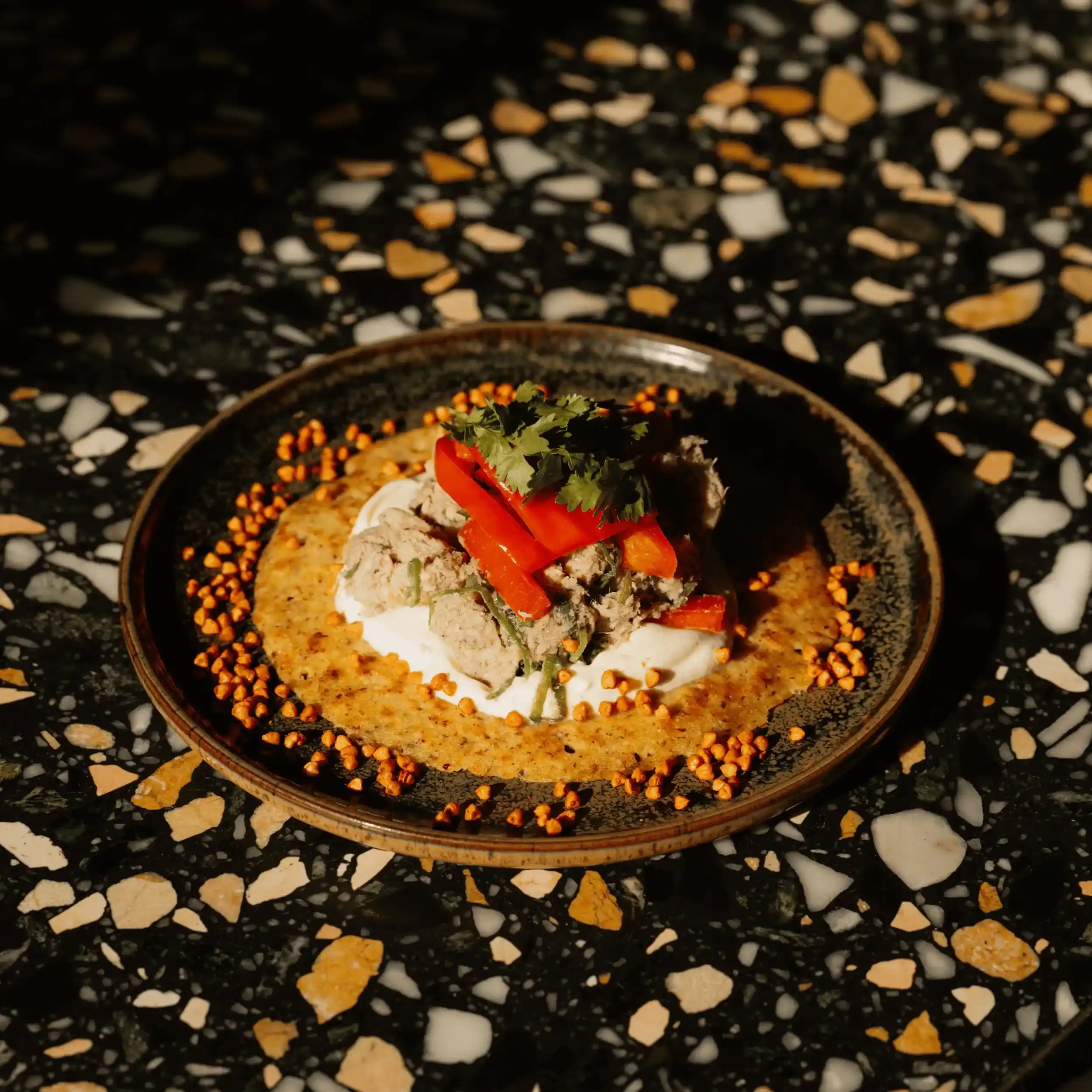 Plato con base cremosa, trozos de atún, tiras de pimiento rojo y guarnición de cilantro sobre una mesa de terrazo.