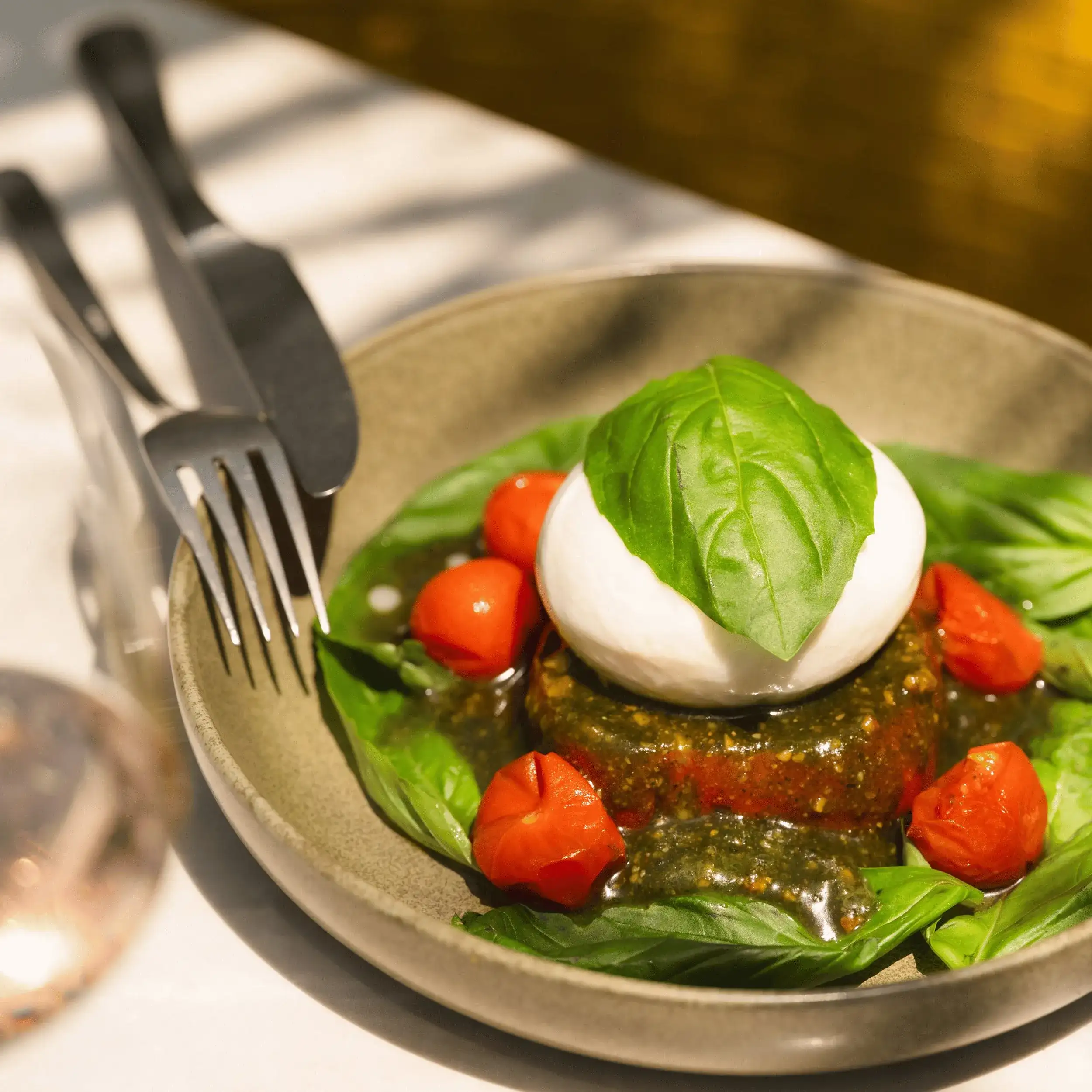 Sirva con mozzarella fresca, hojas de albahaca, salsa pesto y tomates cherry sobre una mesa con cubiertos.