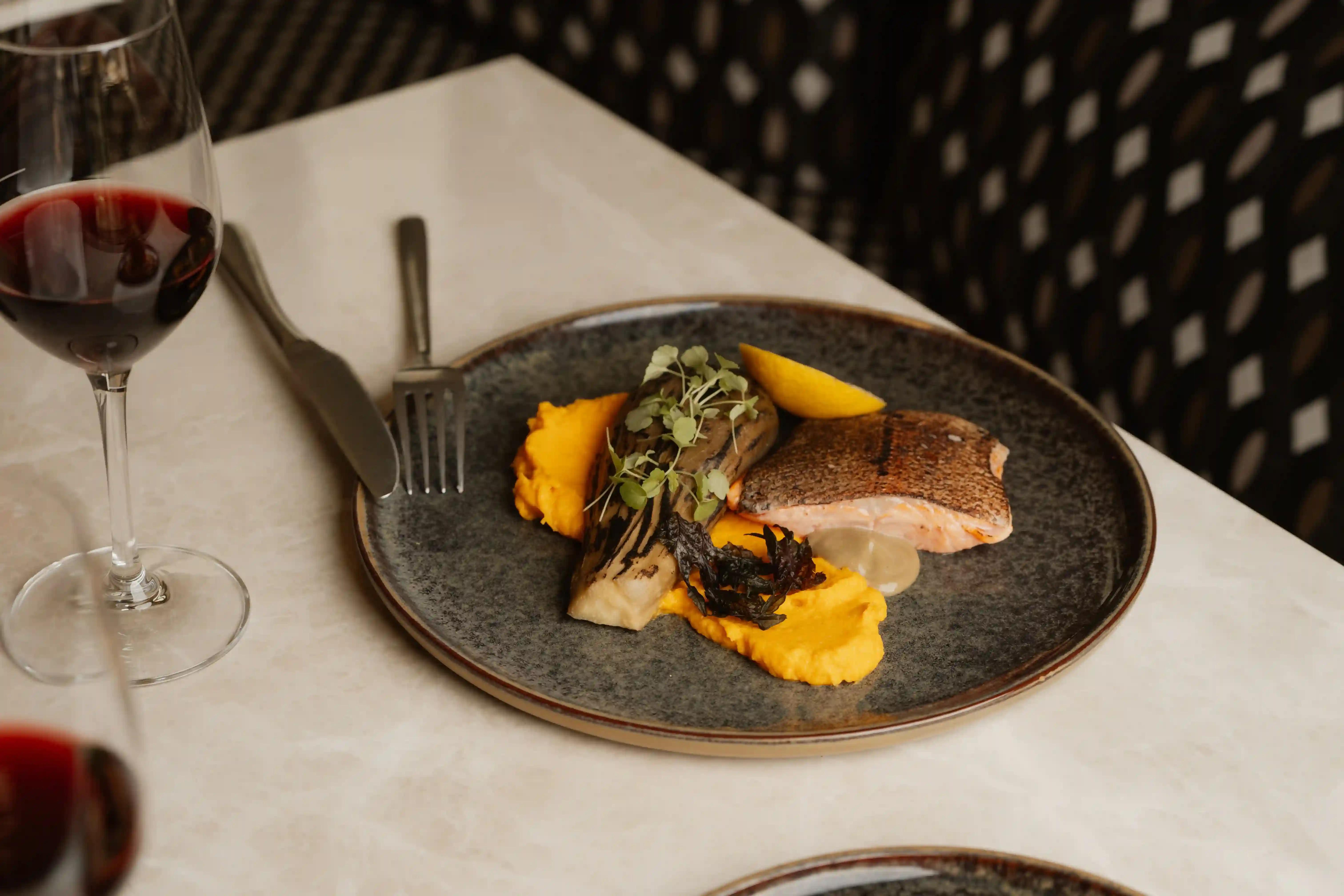 Plato con pescado cocido, puré de verduras de color naranja, berenjenas a la parrilla, verduras, rodajas de limón y una copa de vino tinto.