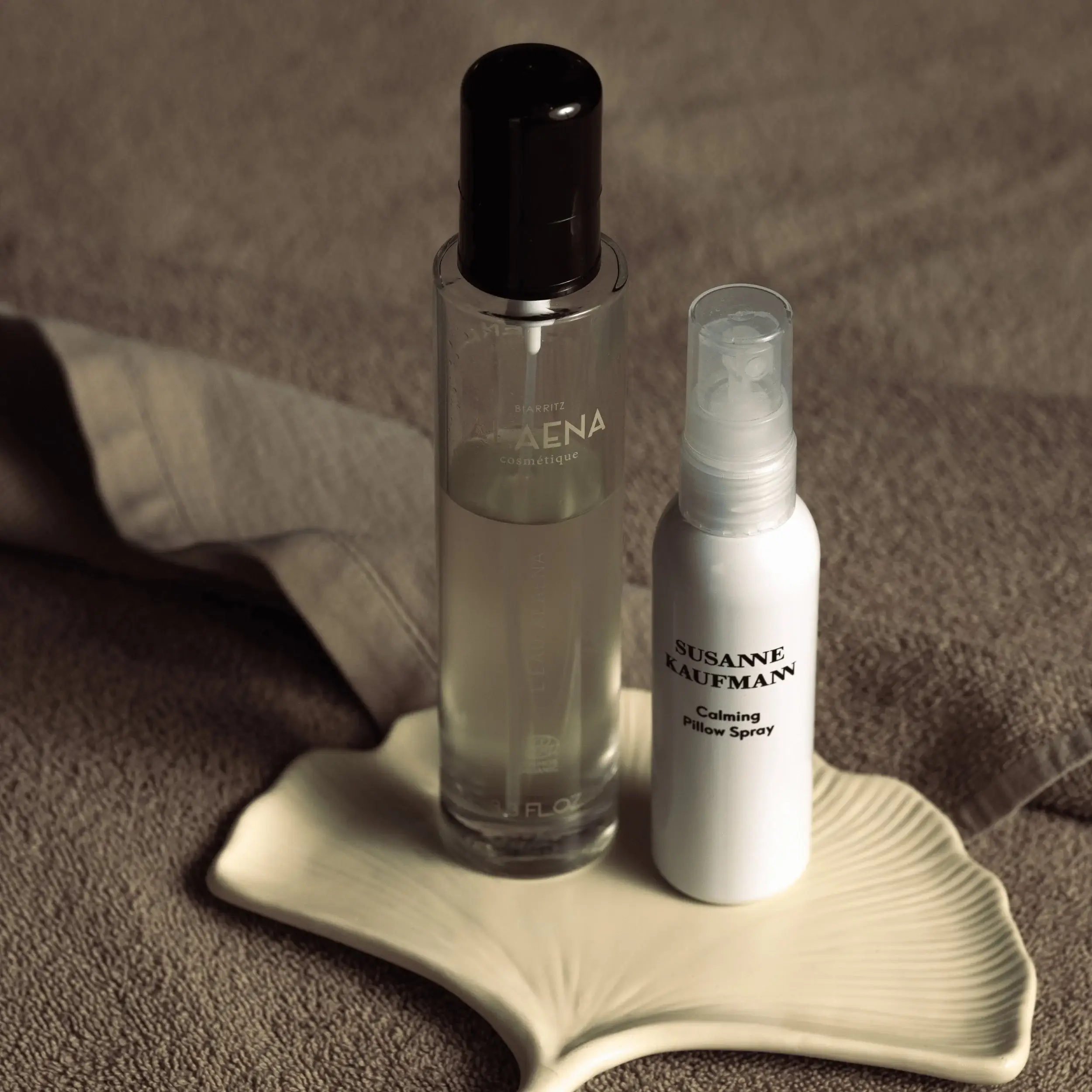 Dos botellas de spray cosmético sobre una tela texturizada de color beige y un plato blanco en forma de hoja.