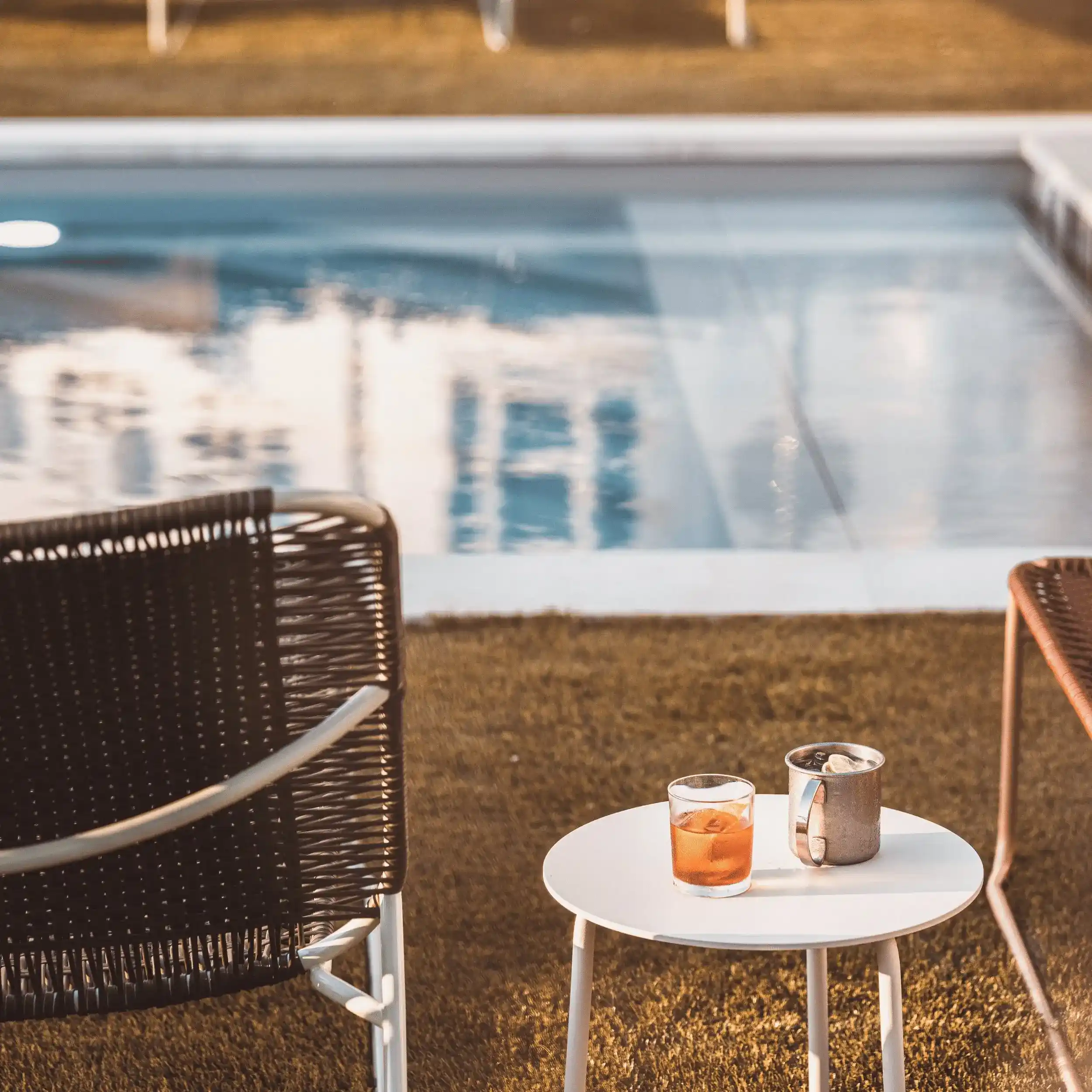 Escena al aire libre con un vaso de bebida de naranja y una taza de metal sobre una mesa blanca junto a la piscina.