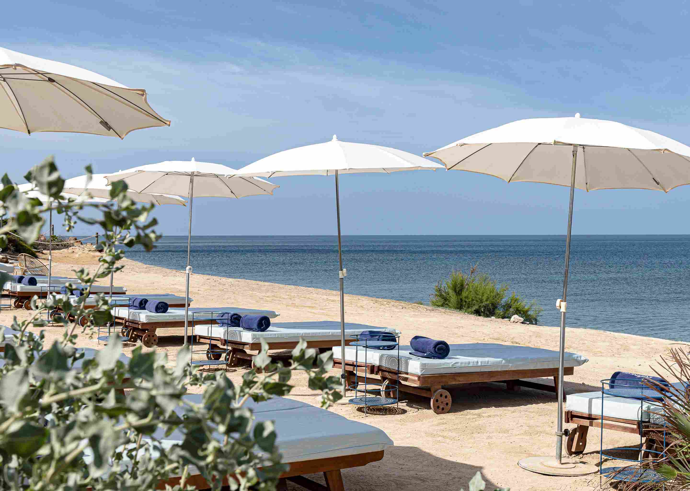 Beach lounge chairs with rolled towels under white umbrellas facing the calm ocean on a sunny day.