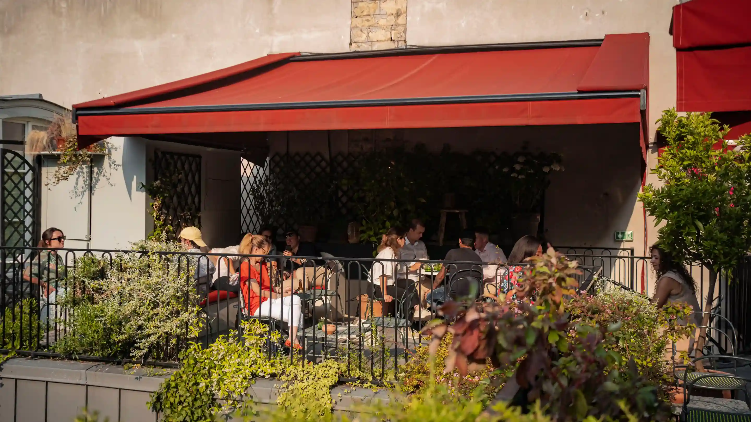 Gente sentada y charlando bajo un toldo rojo en una cafetería con jardín al aire libre con plantas verdes.