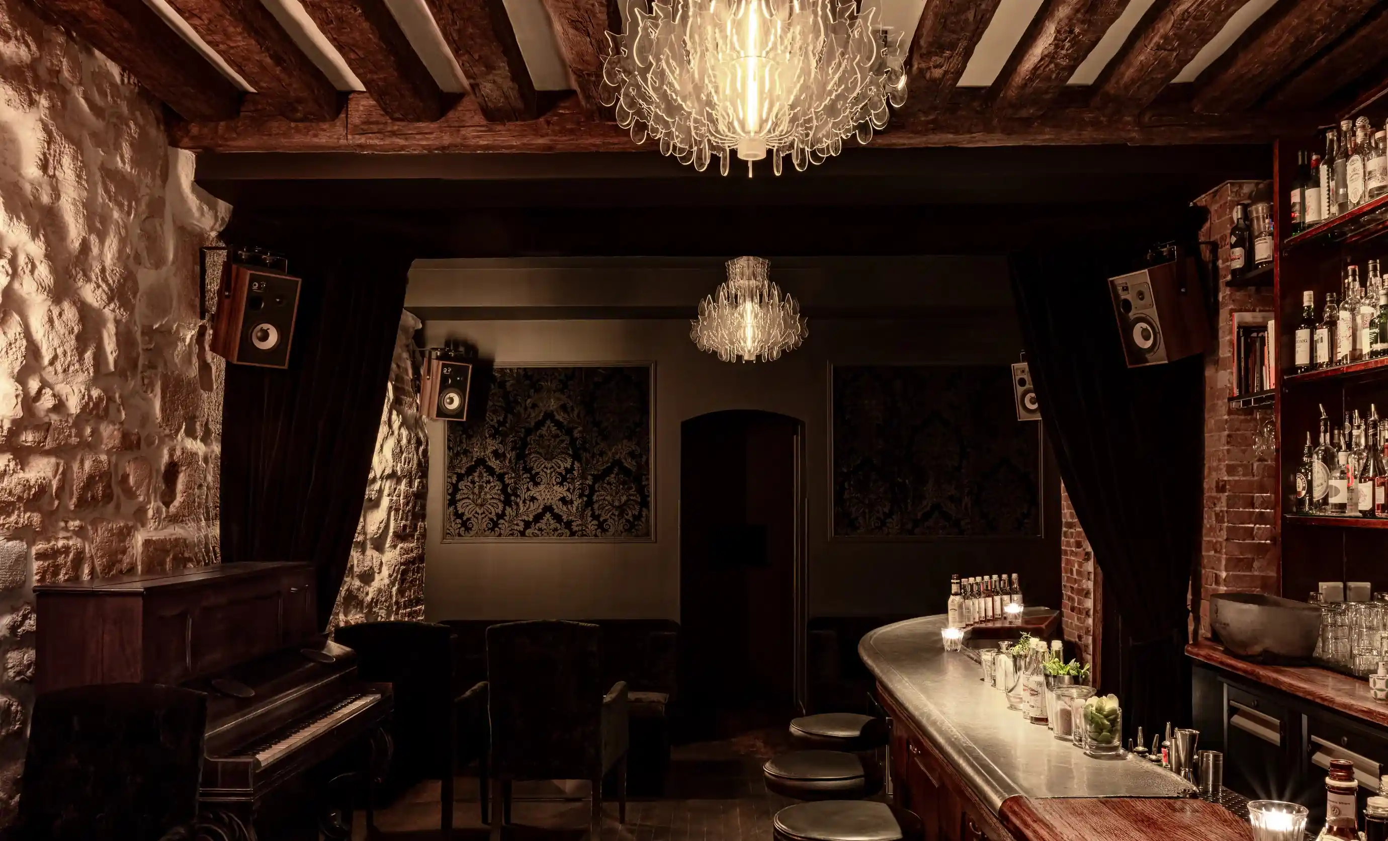 Bar vintage poco illuminato con travi in legno, lampadario, pianoforte, sgabelli da bar e bottiglie sugli scaffali.