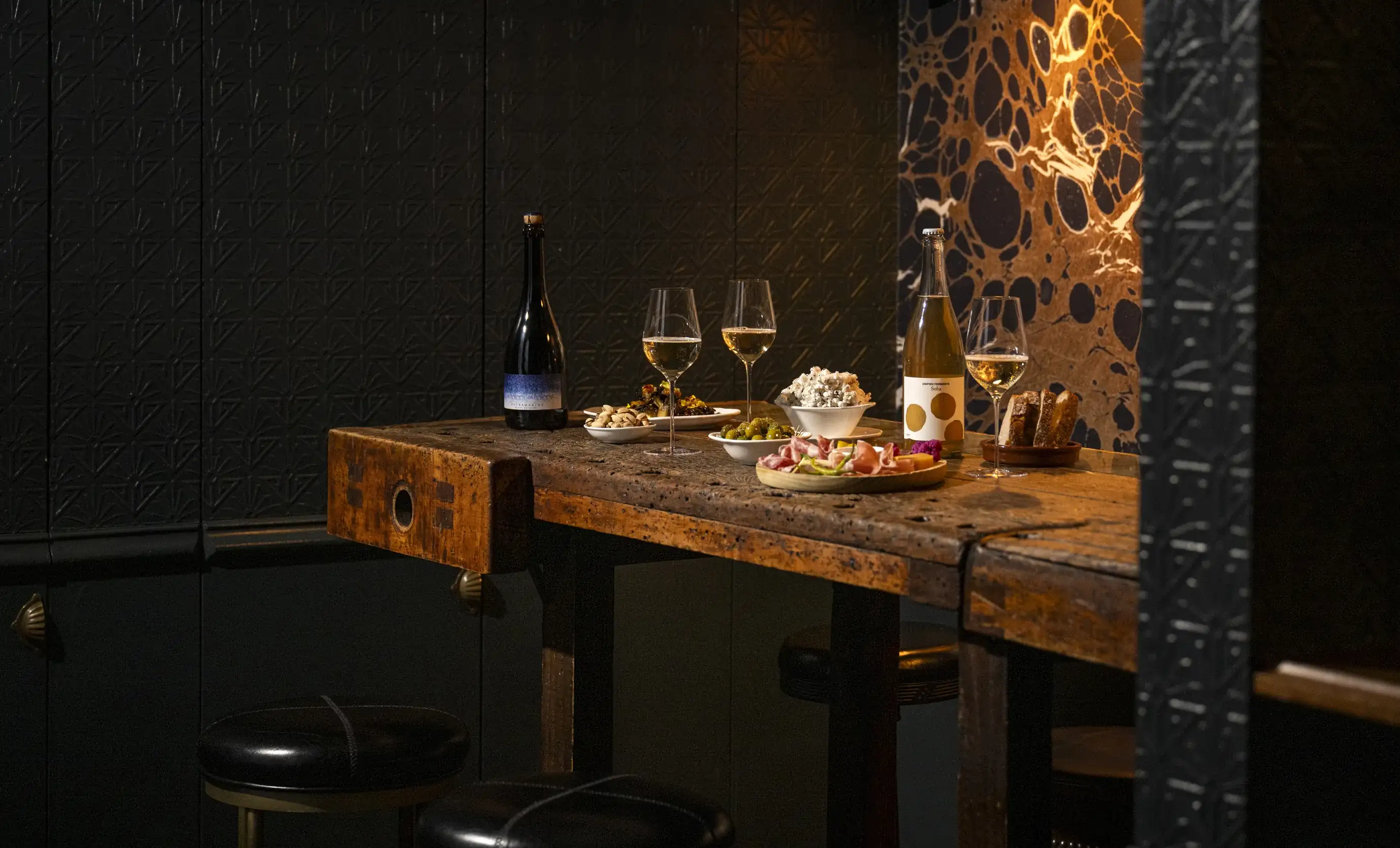 Tavolo rustico in legno con bottiglie di vino, bicchieri, popcorn, noci, olive, pane e salumi in una stanza buia.