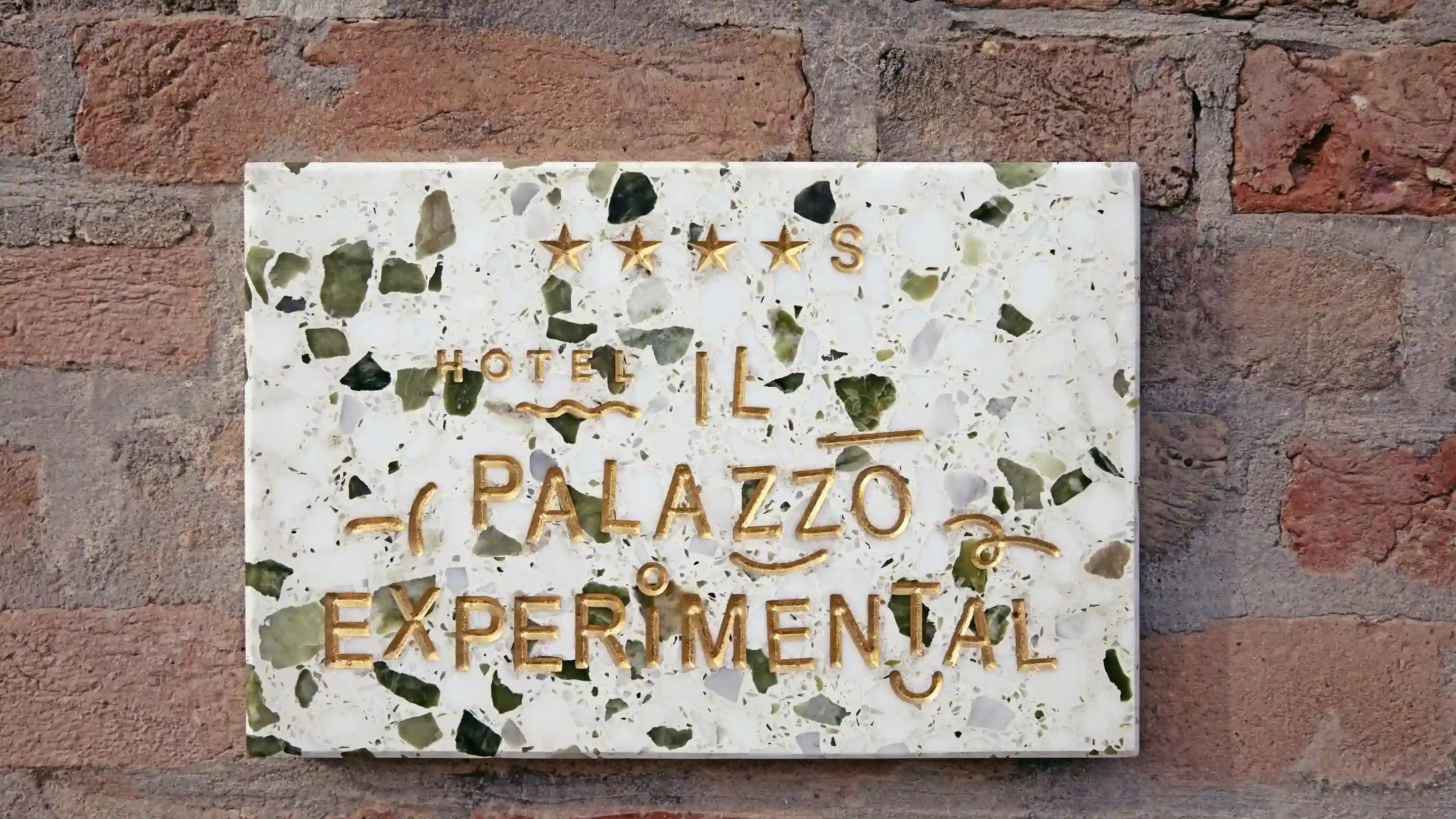 White marble plaque with gold lettering reading 'Hotel Il Palazzo Experimental' on brick wall.