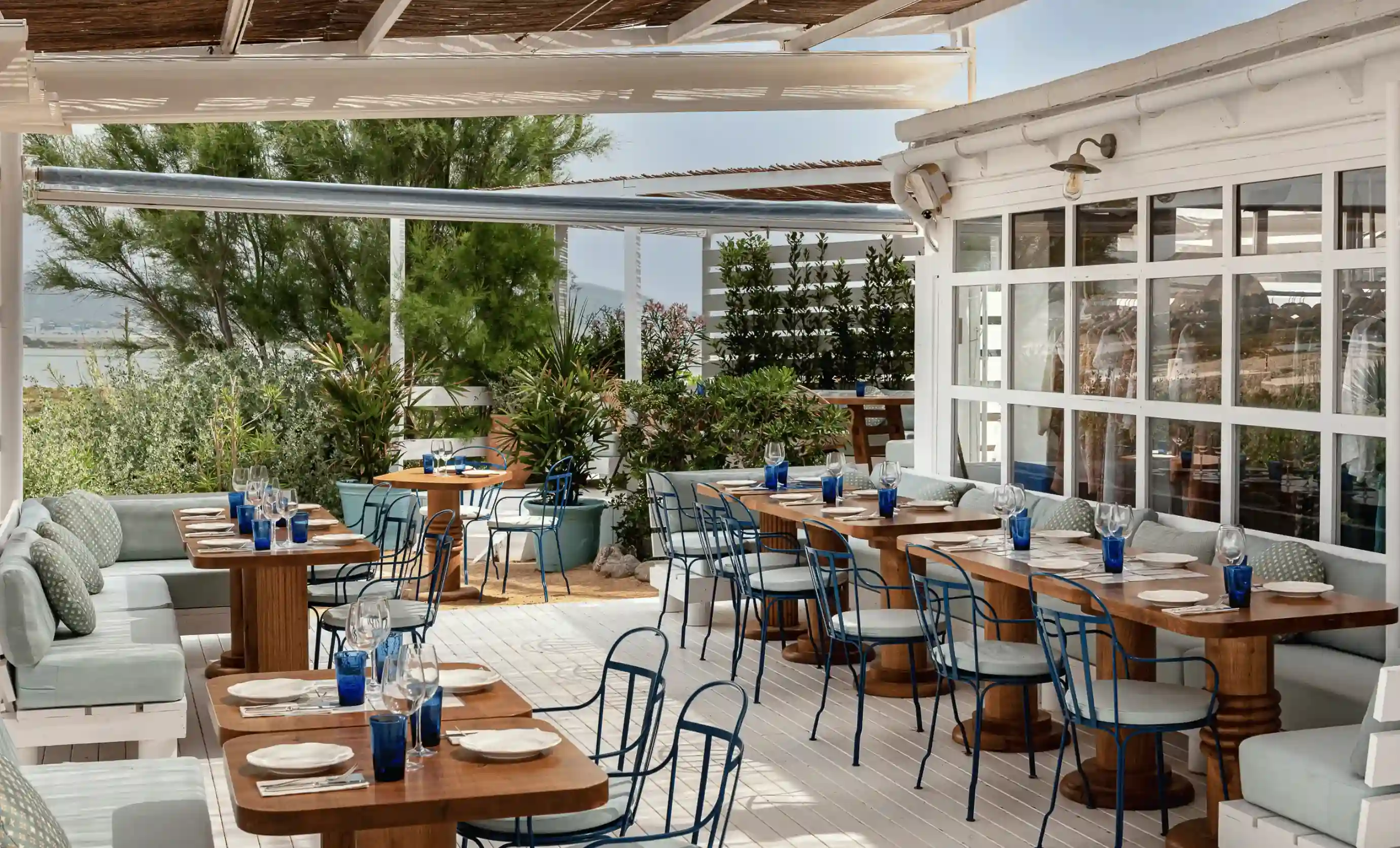 Zona pranzo all'aperto con tavoli in legno, sedie in metallo blu, panche imbottite e vegetazione intorno.