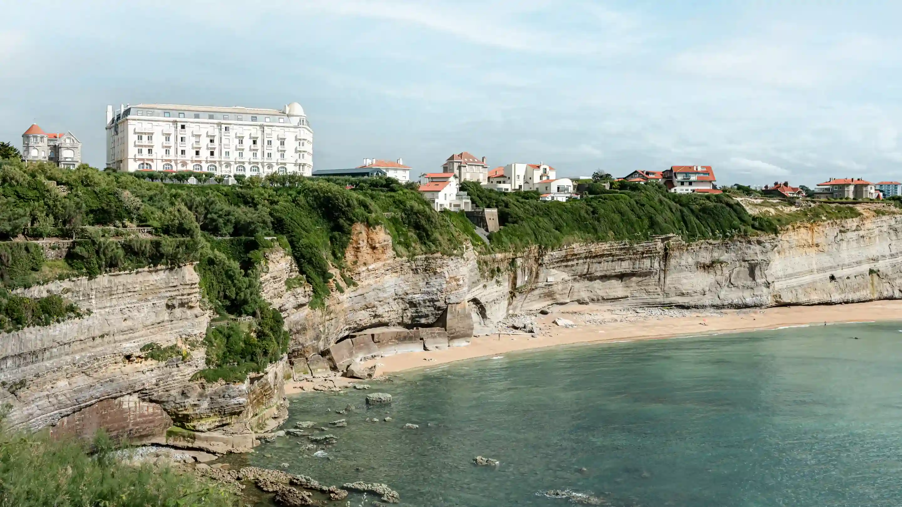 Edifícios brancos em um penhasco verde com vista para um mar azul calmo e uma praia arenosa abaixo.