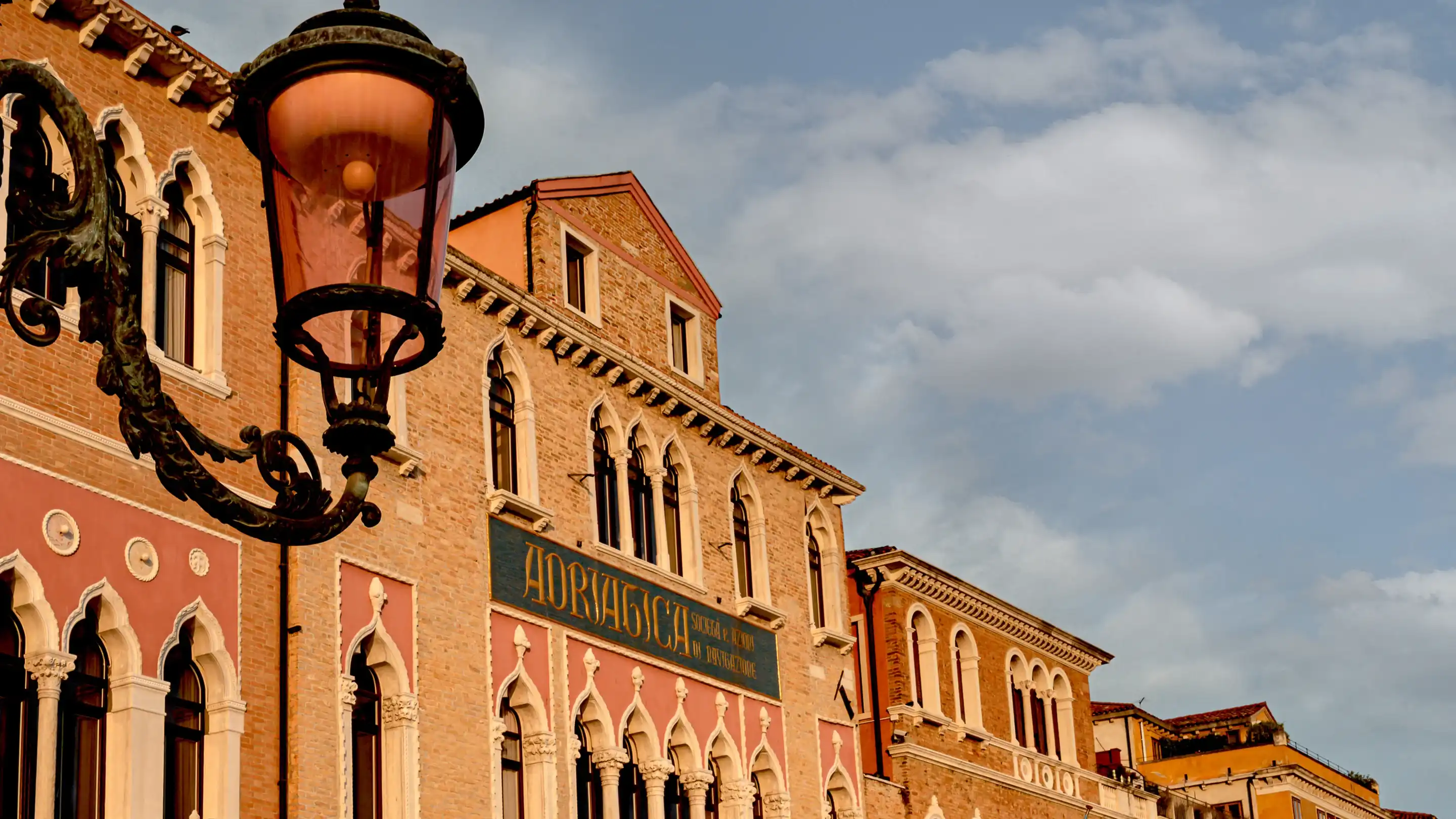 Farola vintage adornada frente a edificios clásicos de estilo veneciano contra un cielo parcialmente nublado.