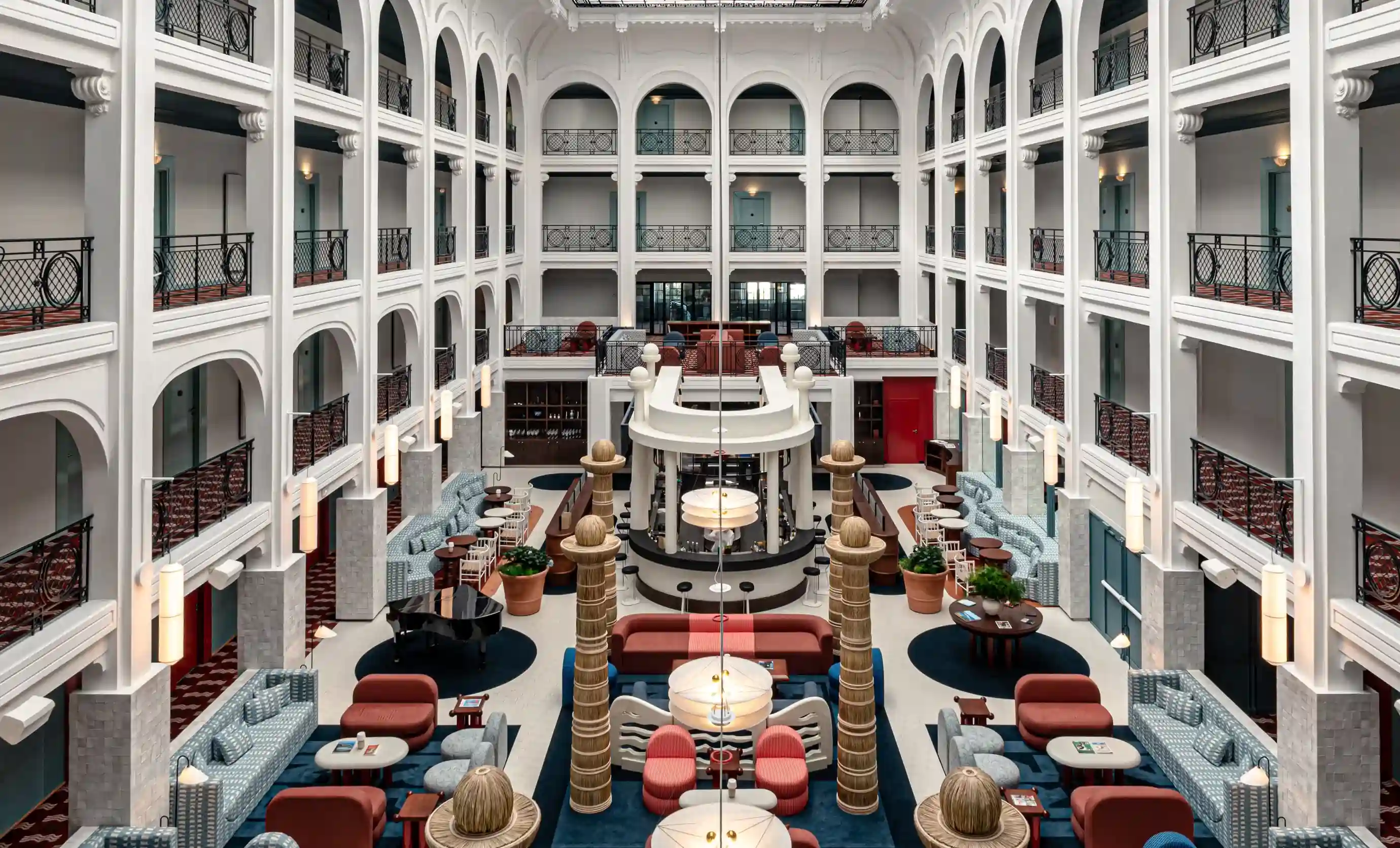 Elegant hotel atrium with white arches, blue and red seating, potted plants, grand piano, and hanging lights.