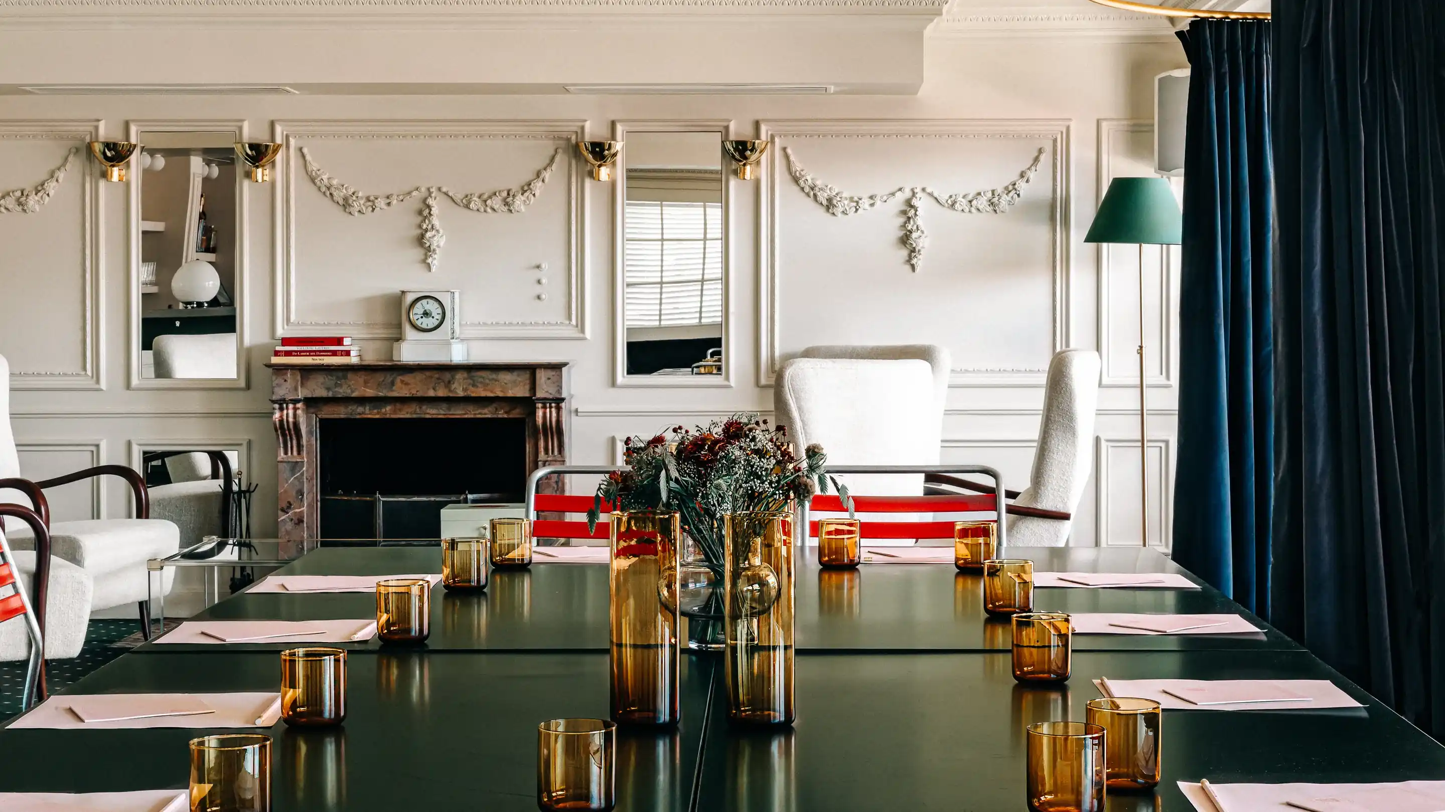 Sala de reuniões elegante com uma mesa grande, copos de âmbar, peça central de flores e um cenário clássico de lareira.