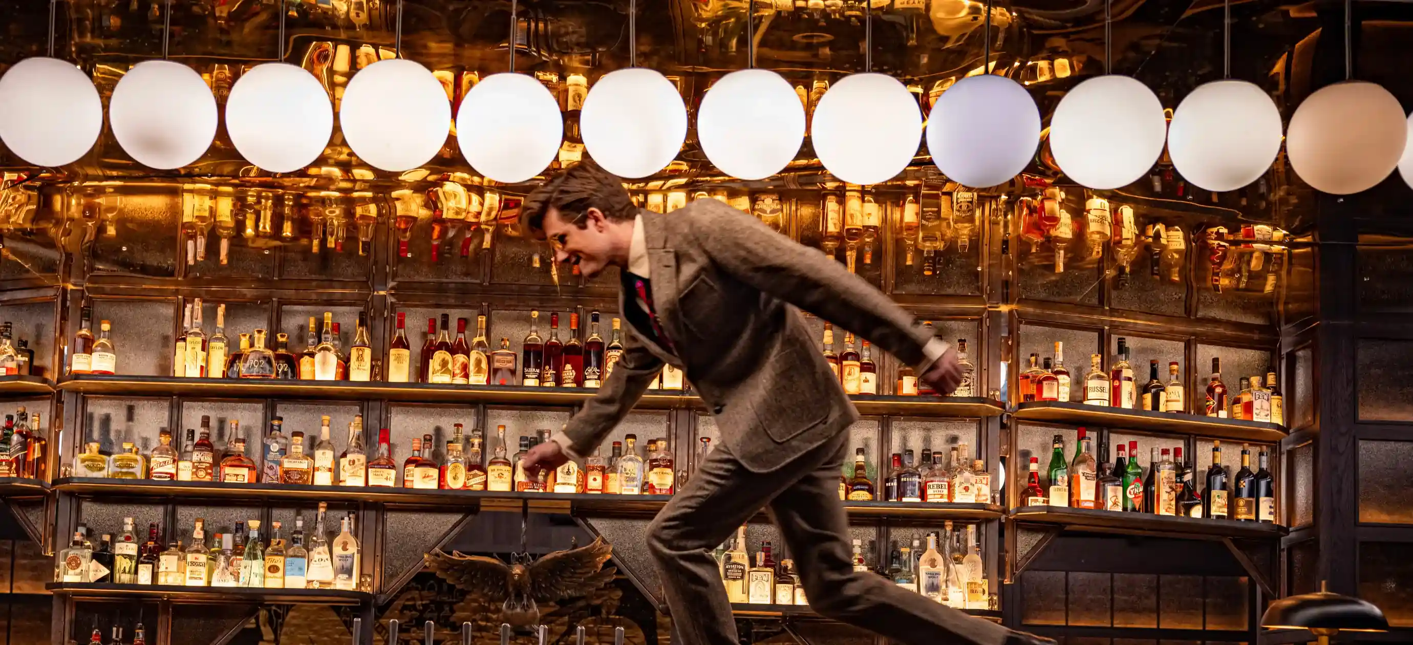 Homme en costume penché en avant dans un bar avec des étagères remplies de bouteilles d'alcool et des lampes rondes suspendues au-dessus.