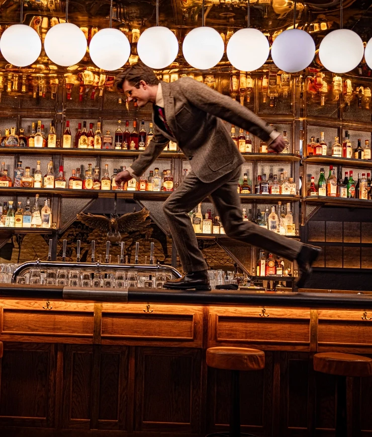 Homme en costume marron courant le long du comptoir d'un bar dans un club faiblement éclairé avec des bouteilles et des lumières derrière.