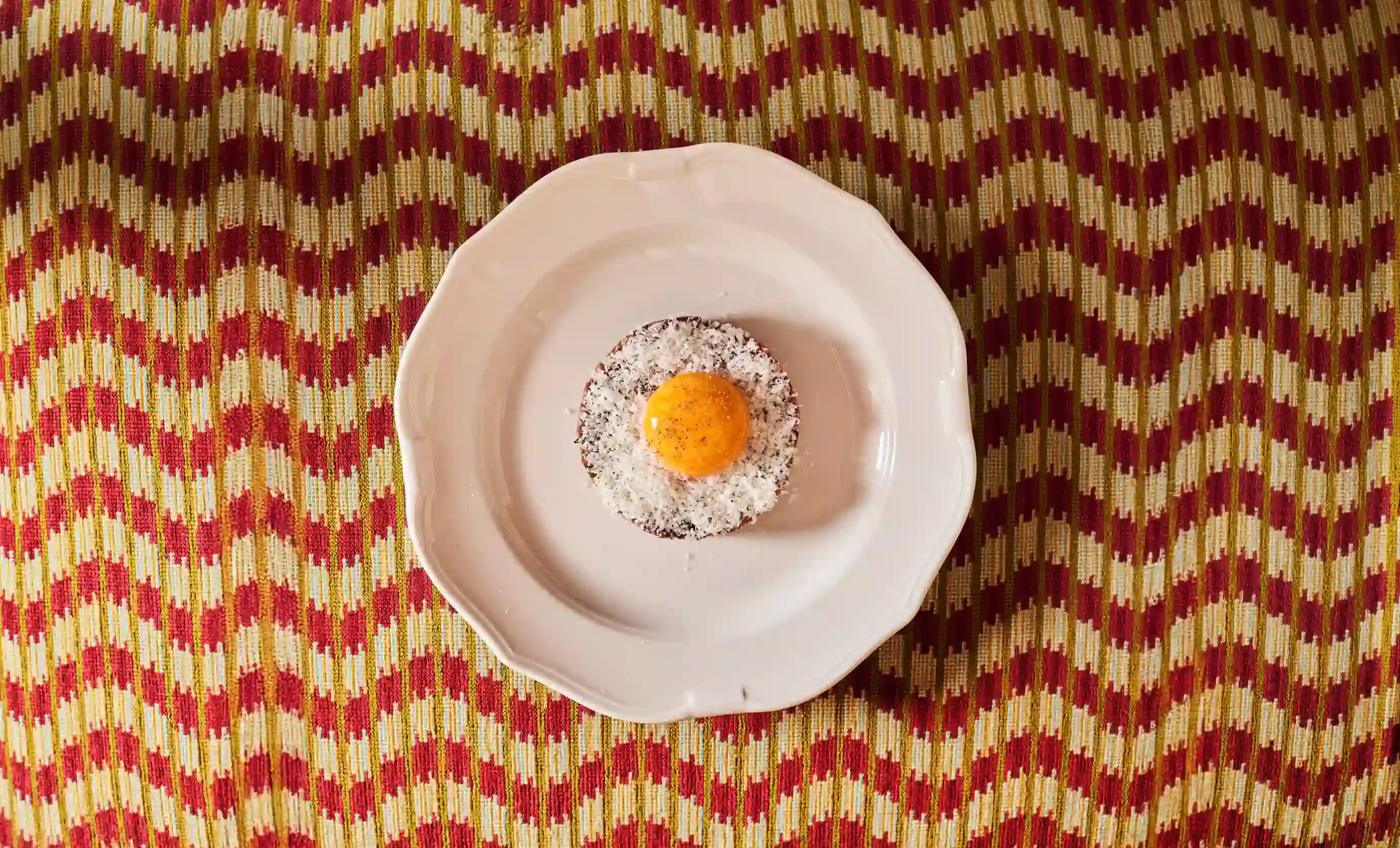 White plate with a dessert topped with orange glaze and white flakes on red-patterned fabric.