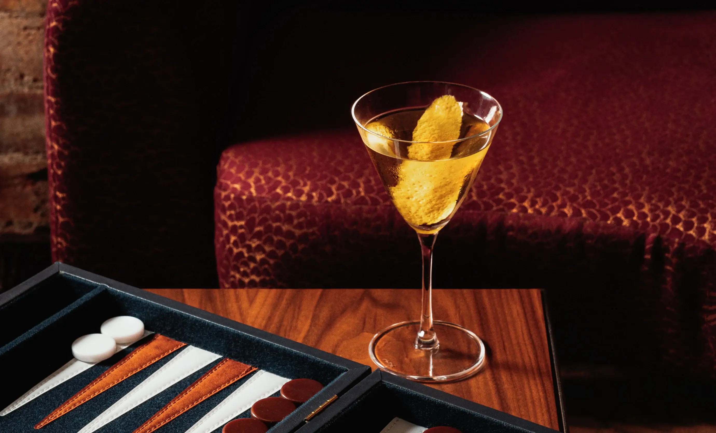 Martini glass with lemon peel on wooden table next to backgammon game with white and brown pieces.