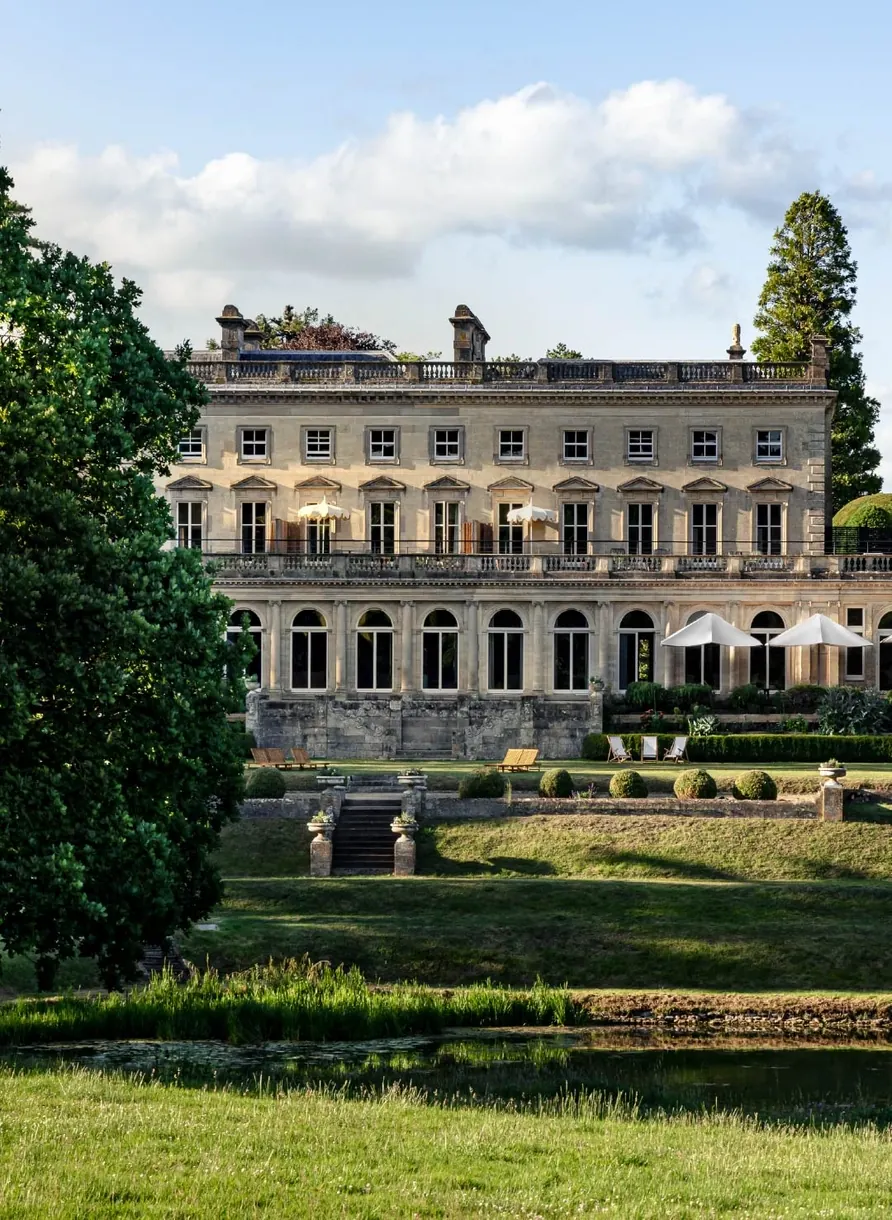 Large historic stone mansion with many windows, lawn chairs, umbrellas, trees, and pond in front.
