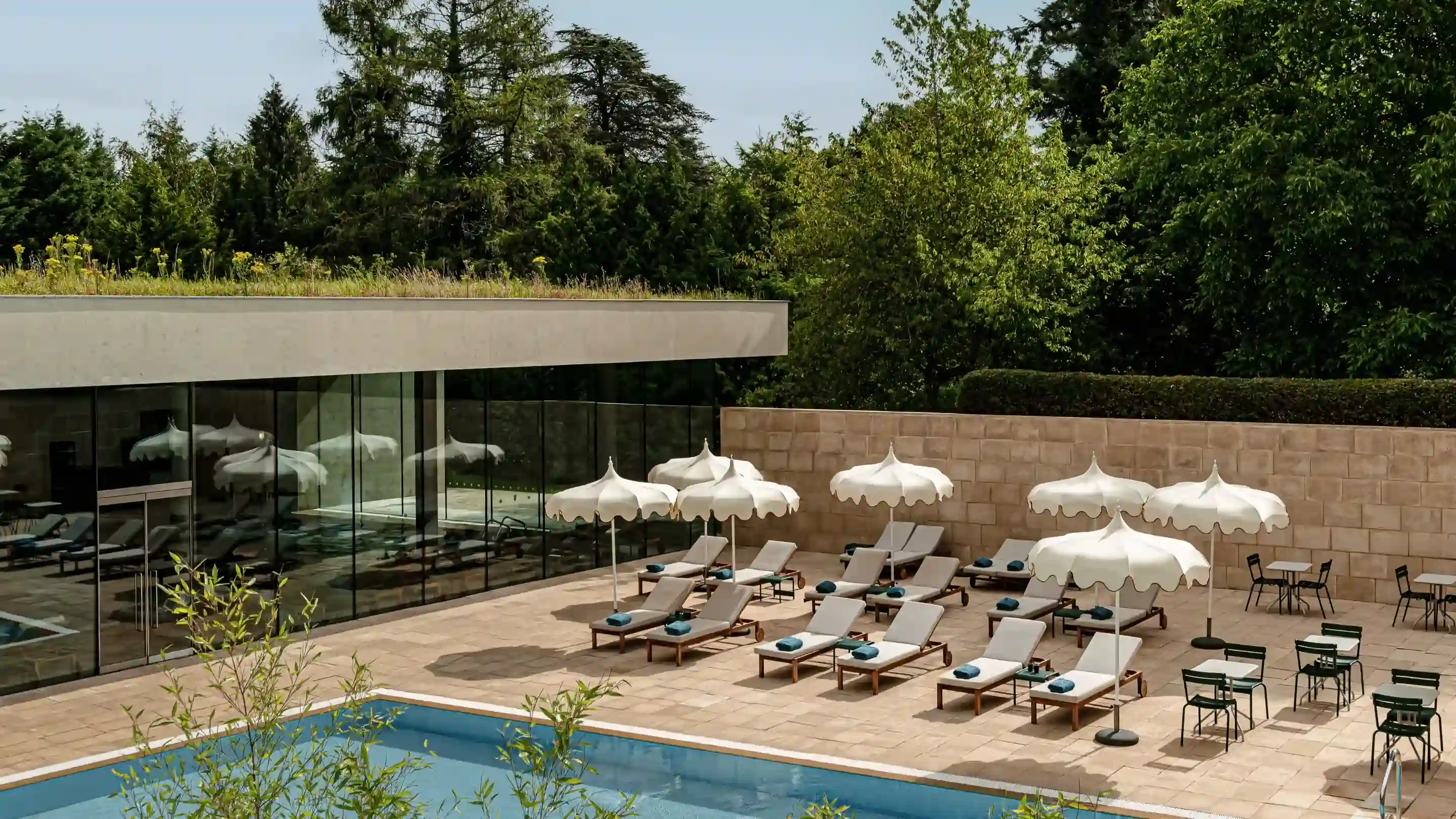 Piscine extérieure avec chaises longues, parasols blancs et petites tables entourées de verdure et de bâtiments en verre.