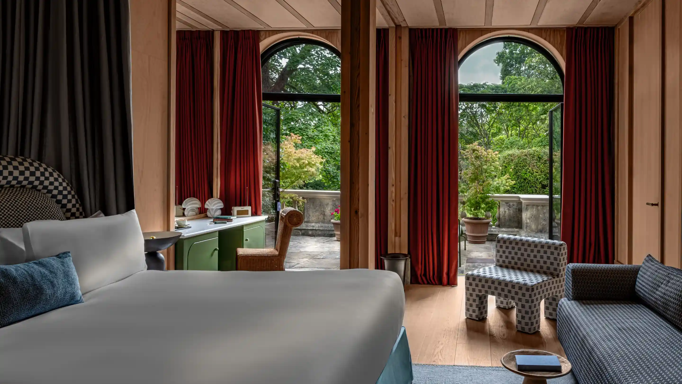 Chambre d'hôtel avec lit blanc, chaise et canapé à motifs, rideaux rouges et portes de balcon ouvertes montrant de la verdure