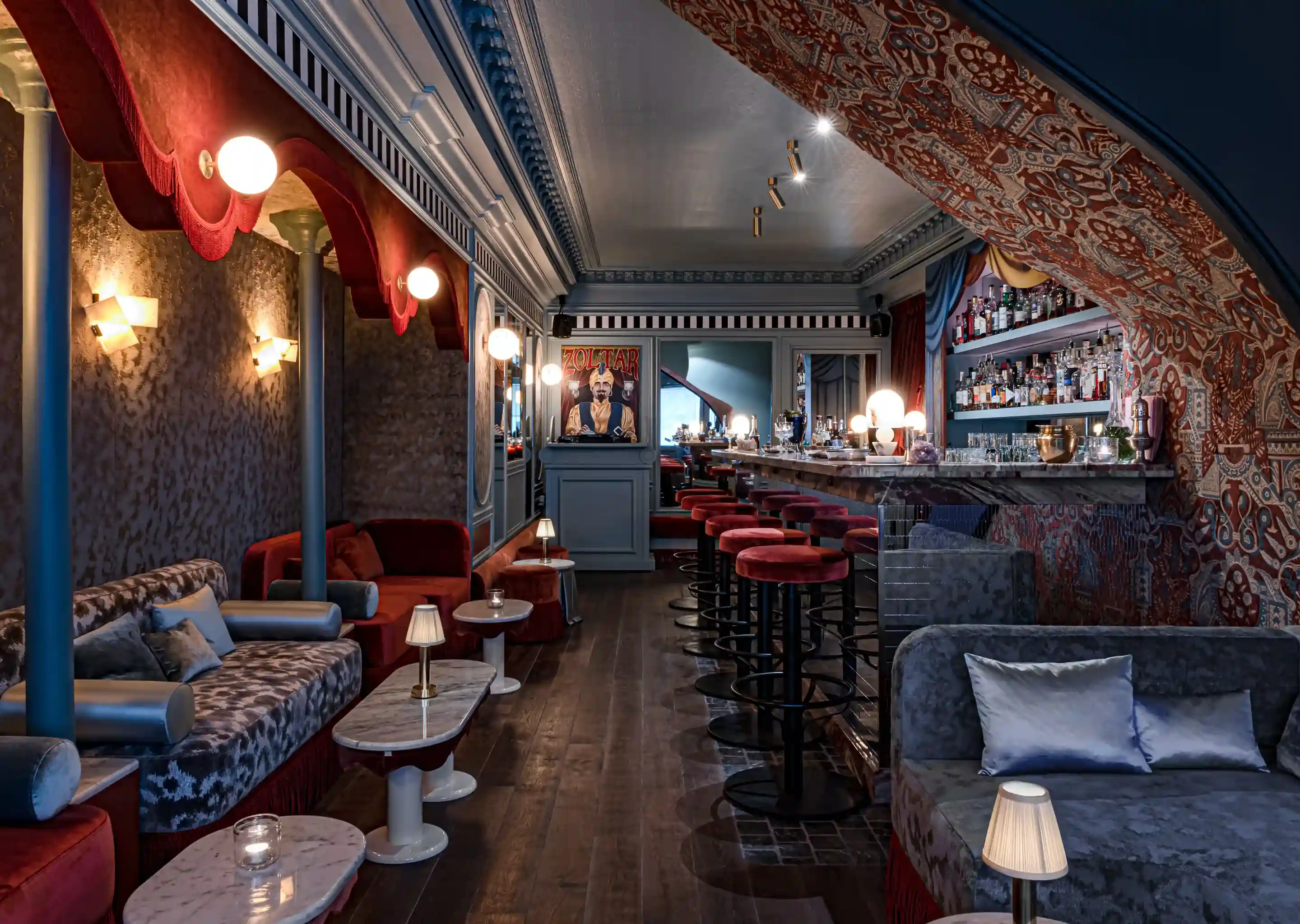 Interno di un elegante bar con tavoli in marmo, sgabelli in velluto rosso, divani e luci calde.