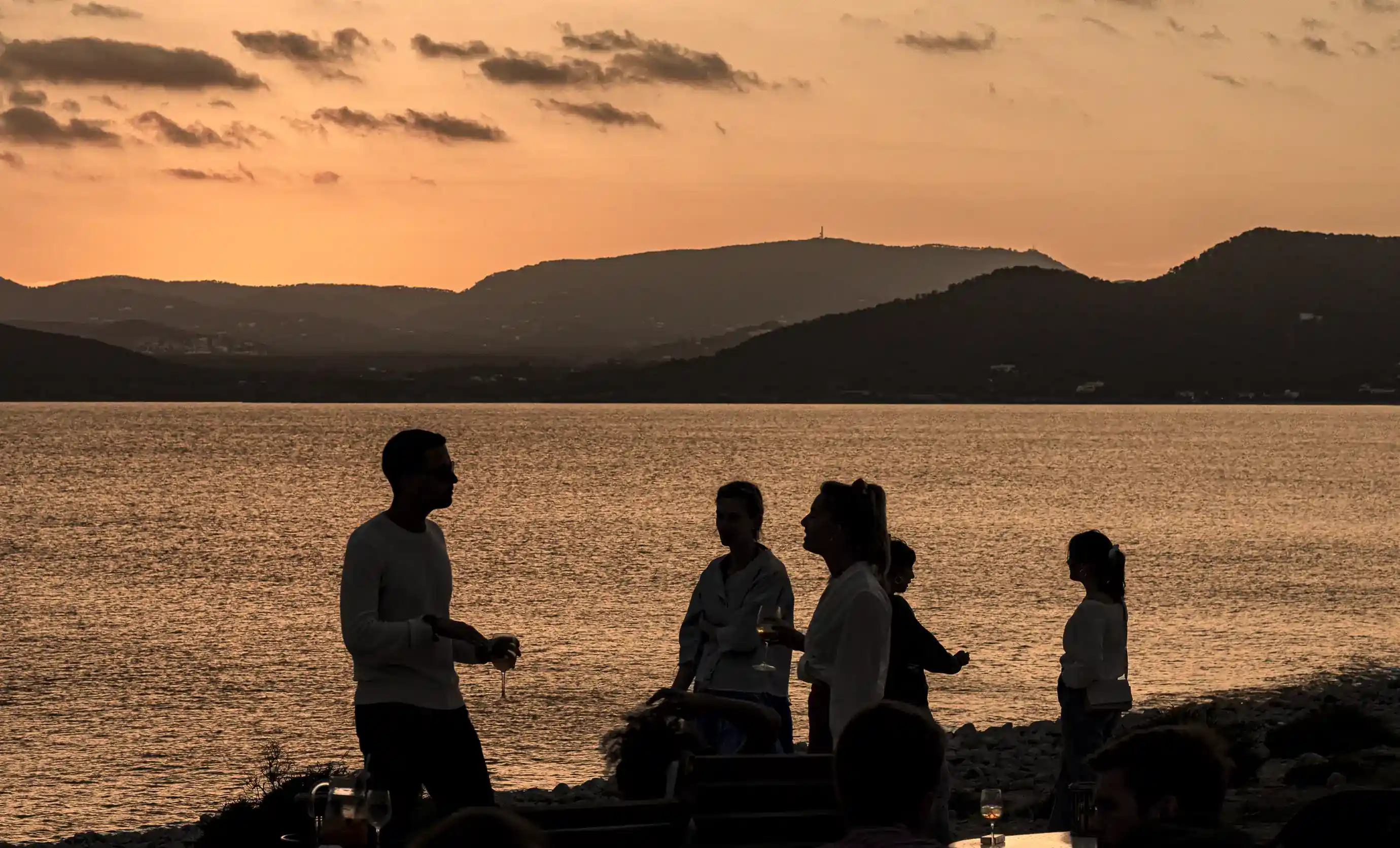 Sagome di persone che socializzano in riva a un lago al tramonto con colline sullo sfondo.