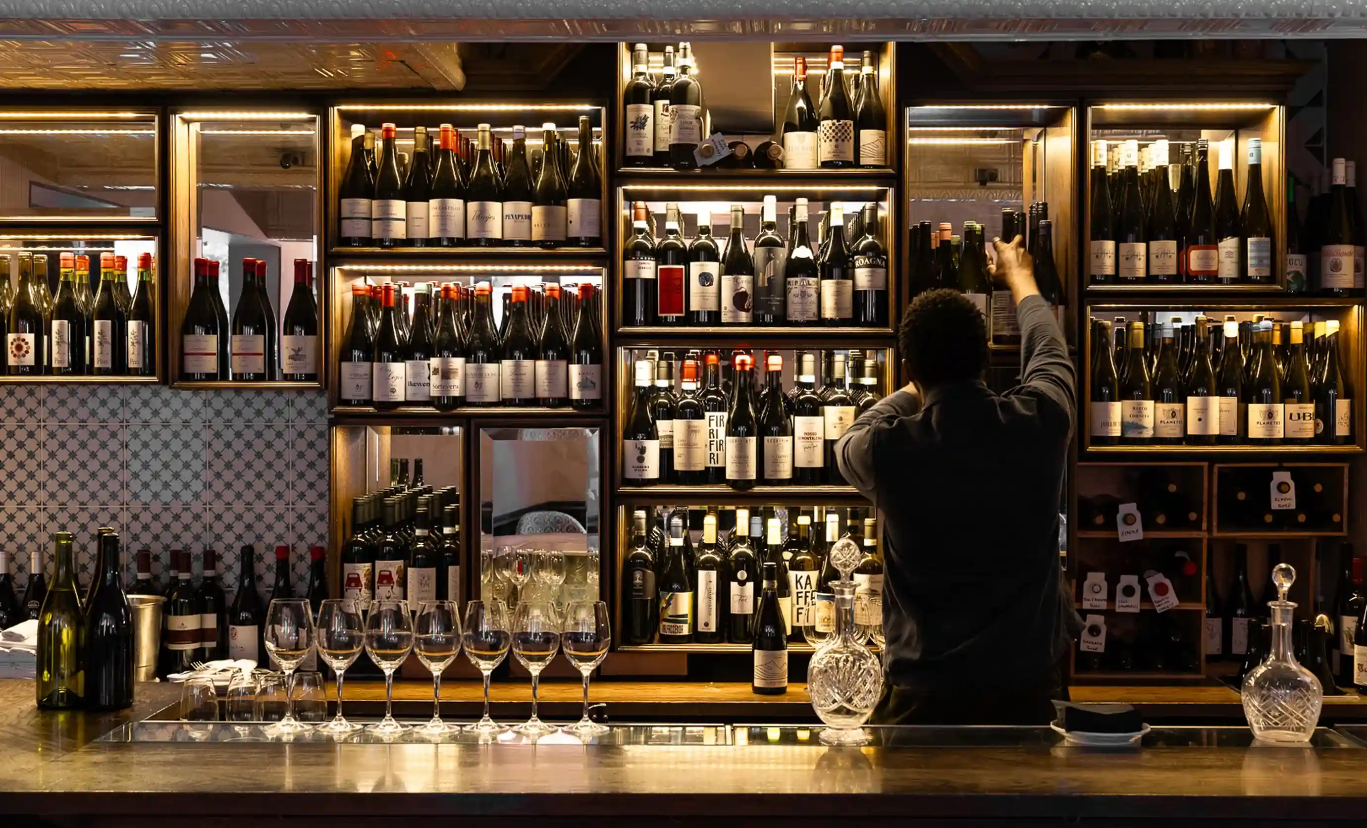 Persona che cerca una bottiglia di vino su uno scaffale da bar retroilluminato pieno di varie bottiglie e bicchieri di vino.