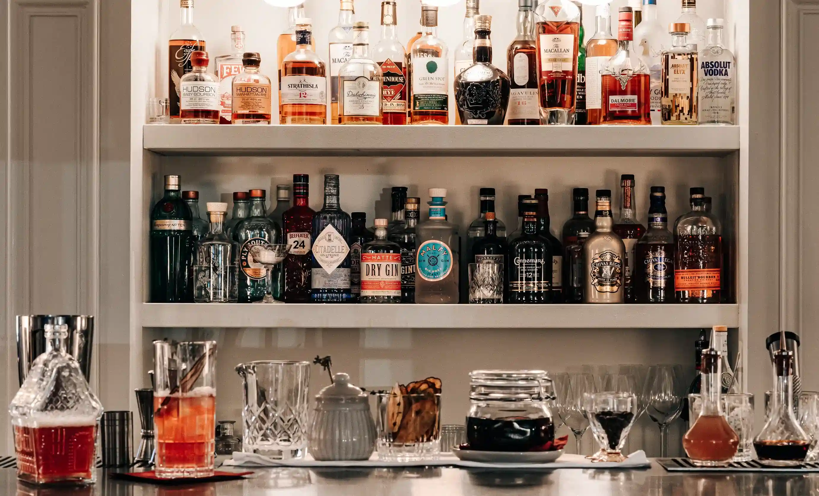Estantes de bar con botellas de licor variadas y una encimera con vasos de cóctel y accesorios para bebidas.