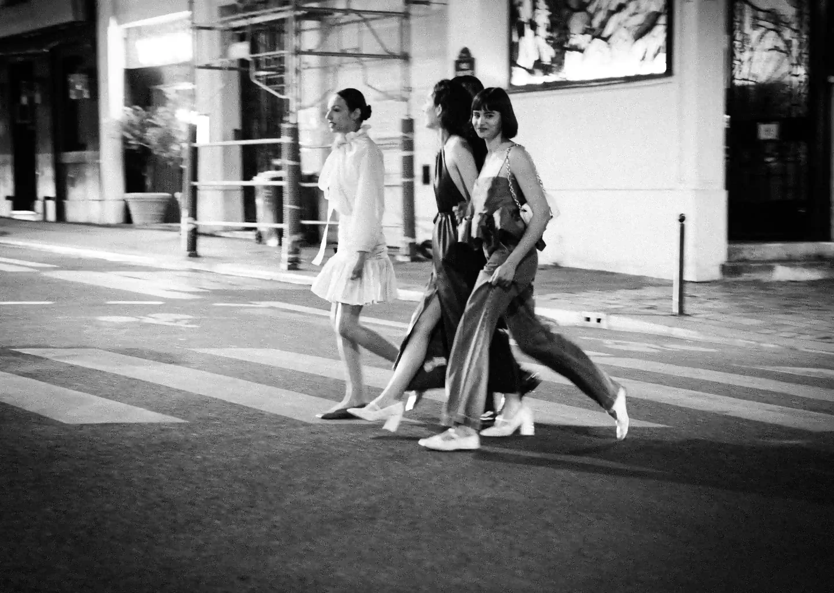 Foto em preto e branco de três mulheres andando por uma rua da cidade em uma faixa de pedestres à noite.