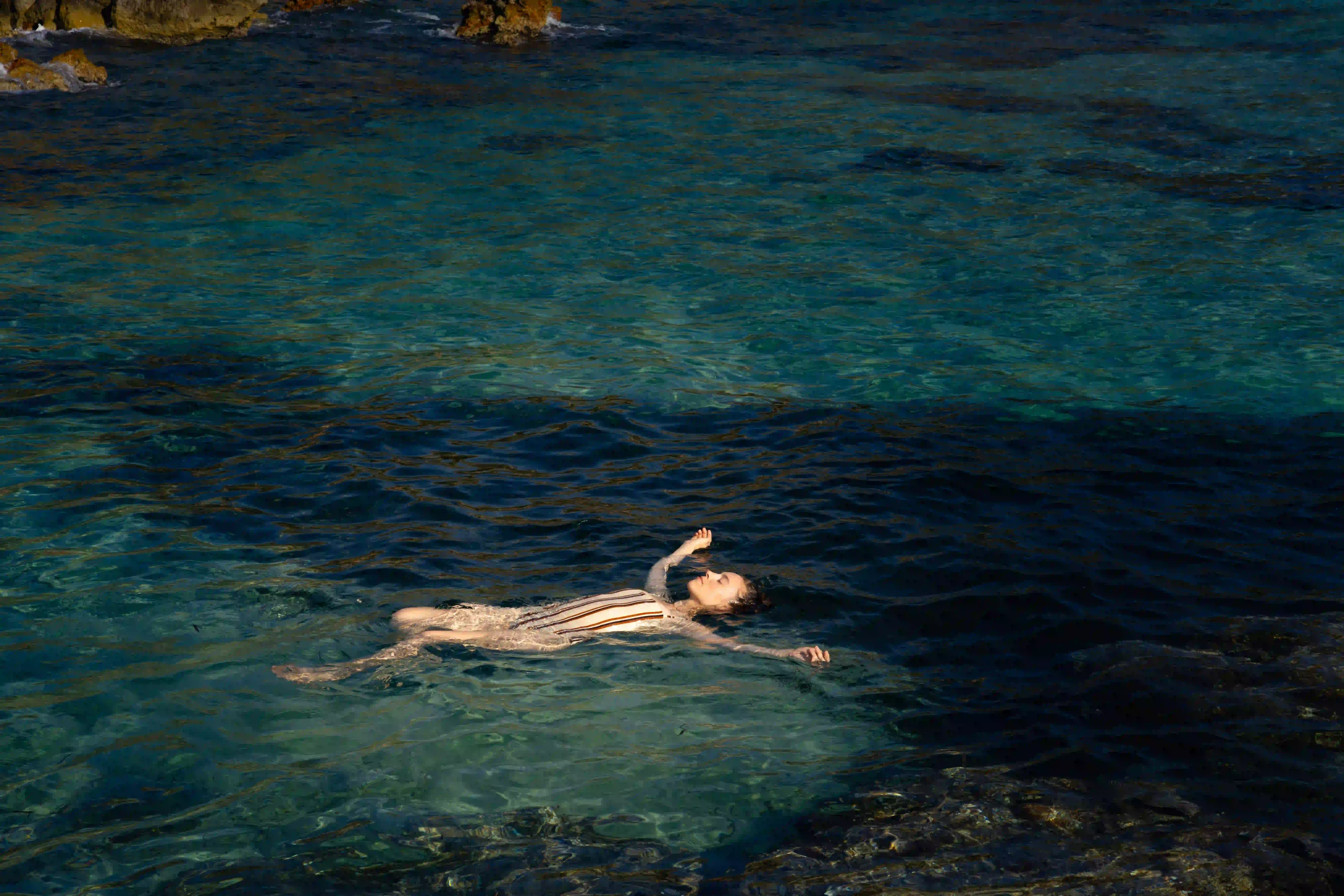 Pessoa flutuando de costas em águas azuis claras perto da costa rochosa.