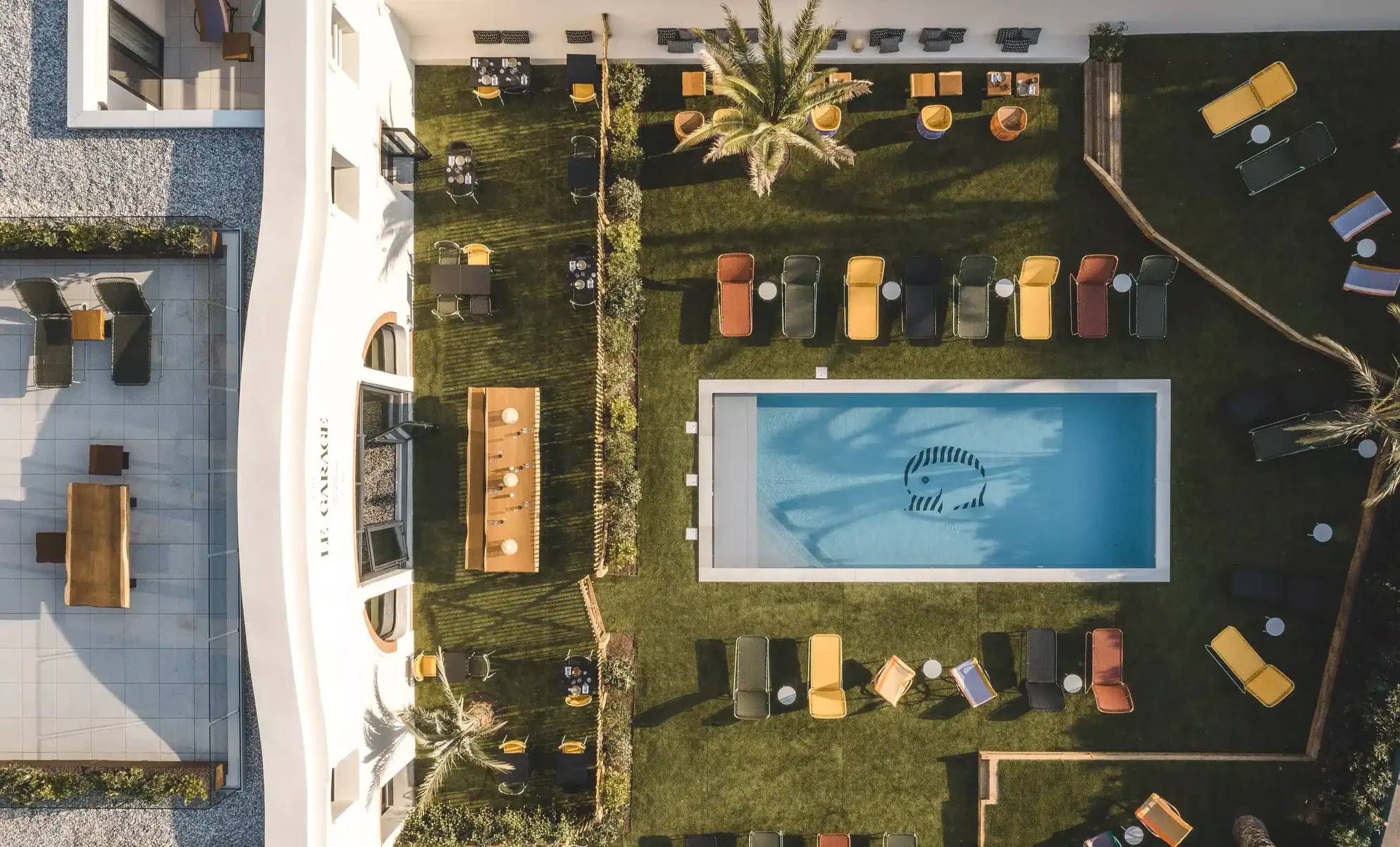 Veduta aerea di una piscina rettangolare con un disegno di pesci, circondata da sedie a sdraio colorate e palme.