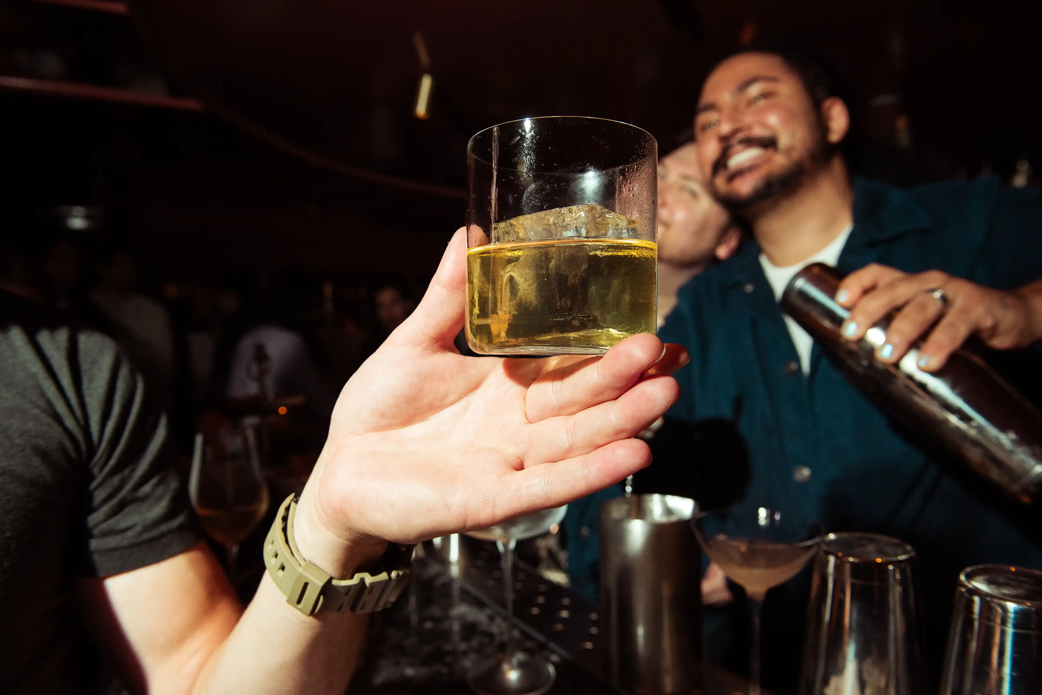Mão segurando um copo com uísque e gelo em um bar com um barman sorridente ao fundo.