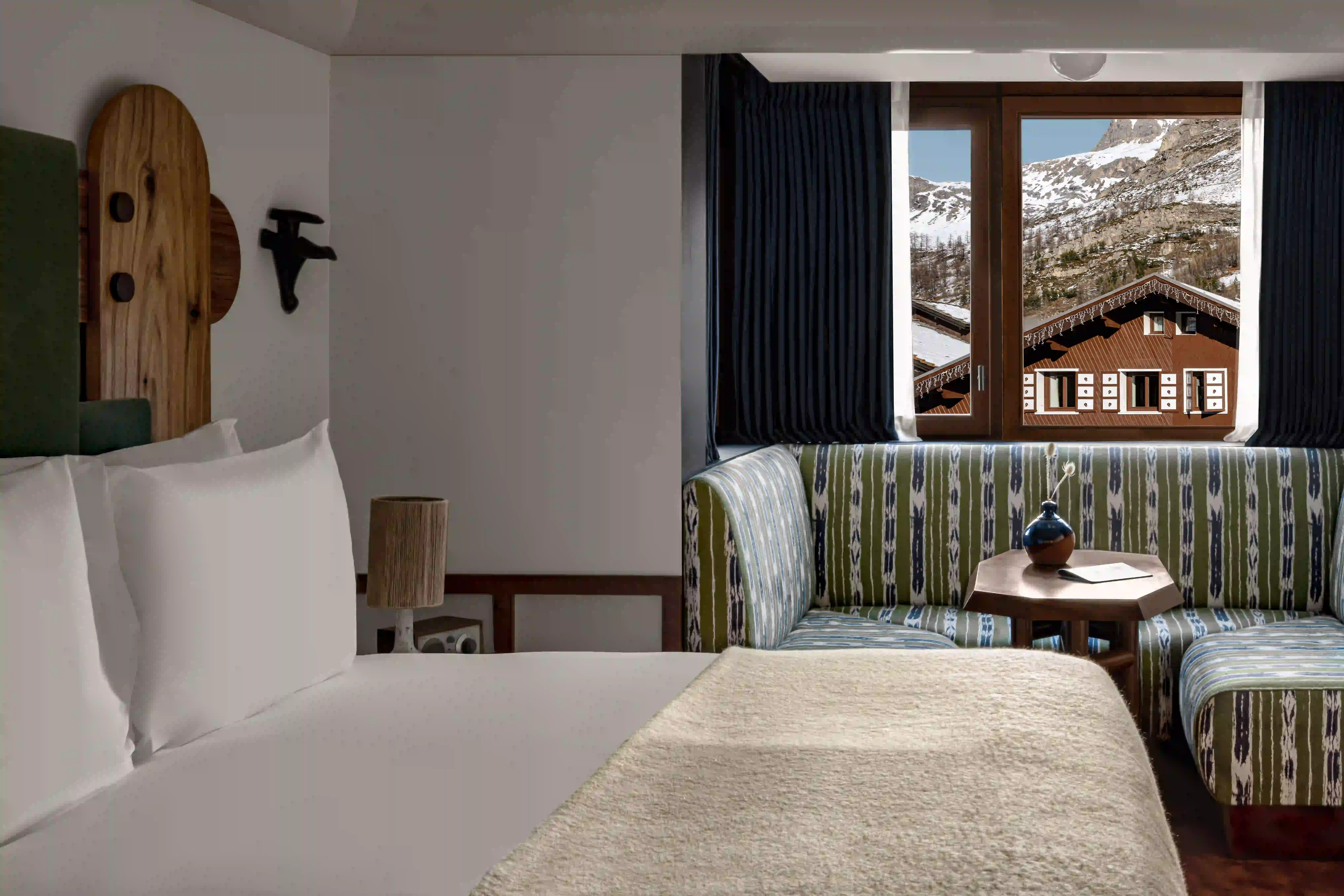 Camera d'albergo con lenzuola bianche e finestra con vista sulla montagna innevata e sul tetto dello chalet.