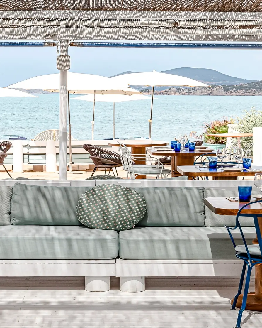 Outdoor restaurant seating with light gray couch, tables, white umbrellas, and ocean view with distant hills.