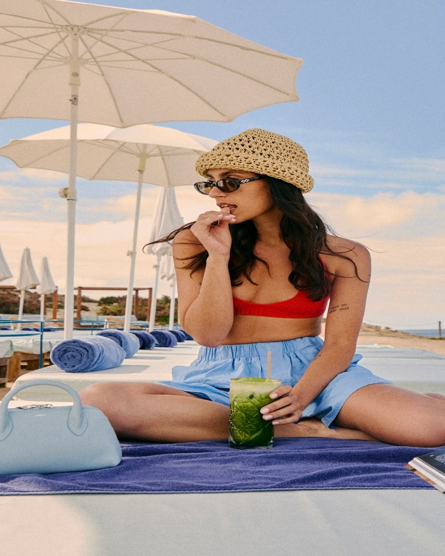 Woman in red bikini top and blue shorts sitting cross-legged on beach chair with drink and umbrellas.