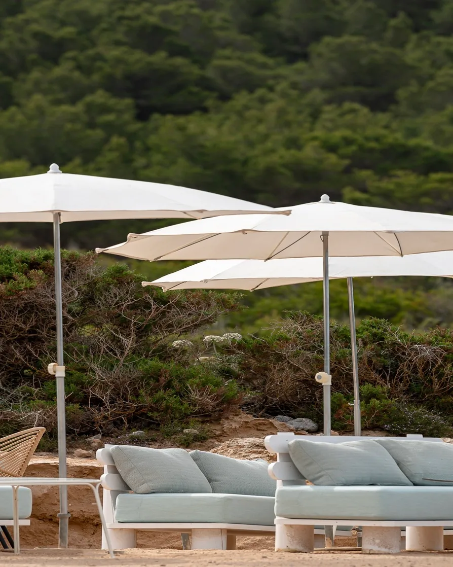 Outdoor seating with light blue cushions under white umbrellas on sandy ground near green bushes.