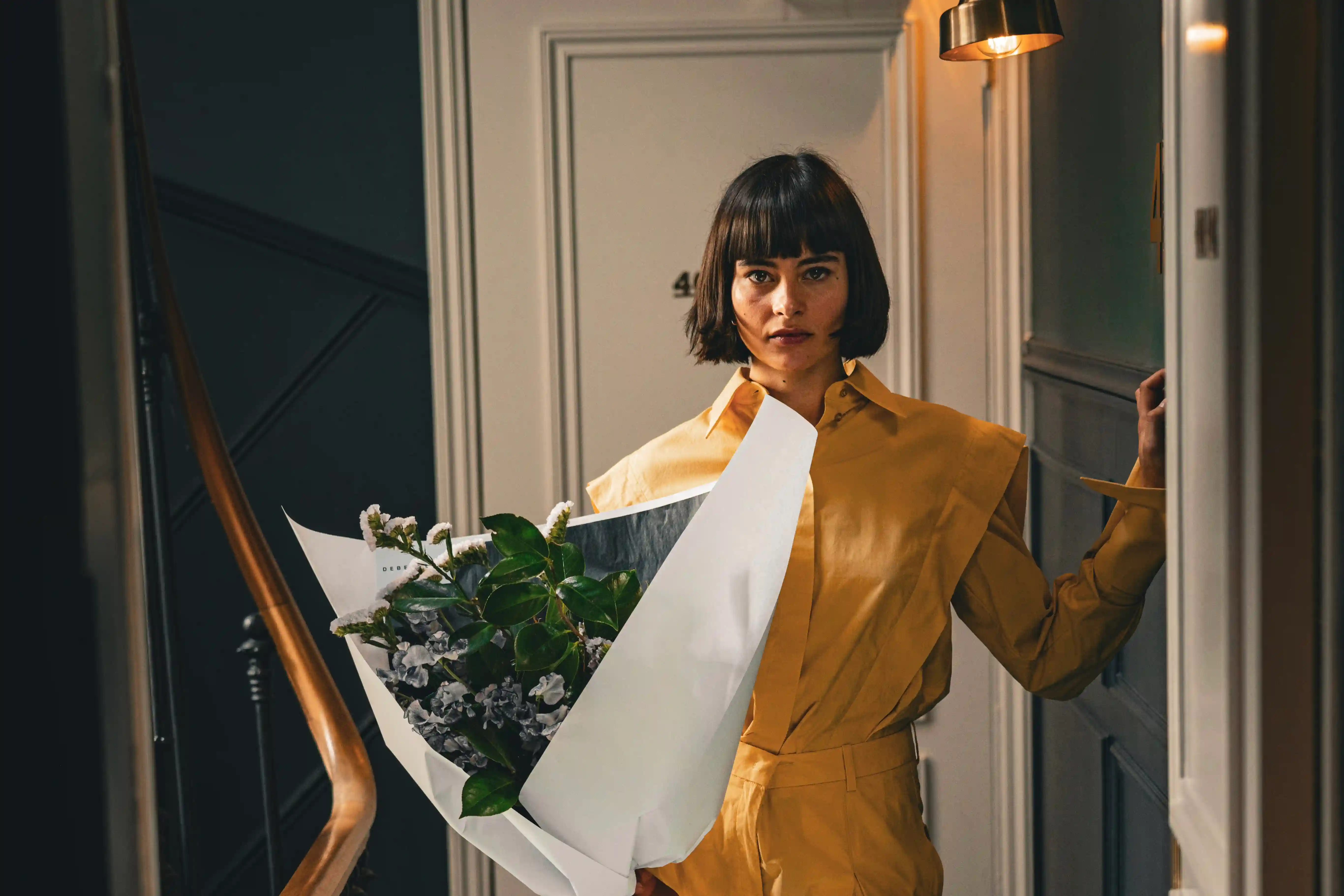 Woman in mustard yellow outfit holding a bouquet of flowers in a dimly lit hallway.