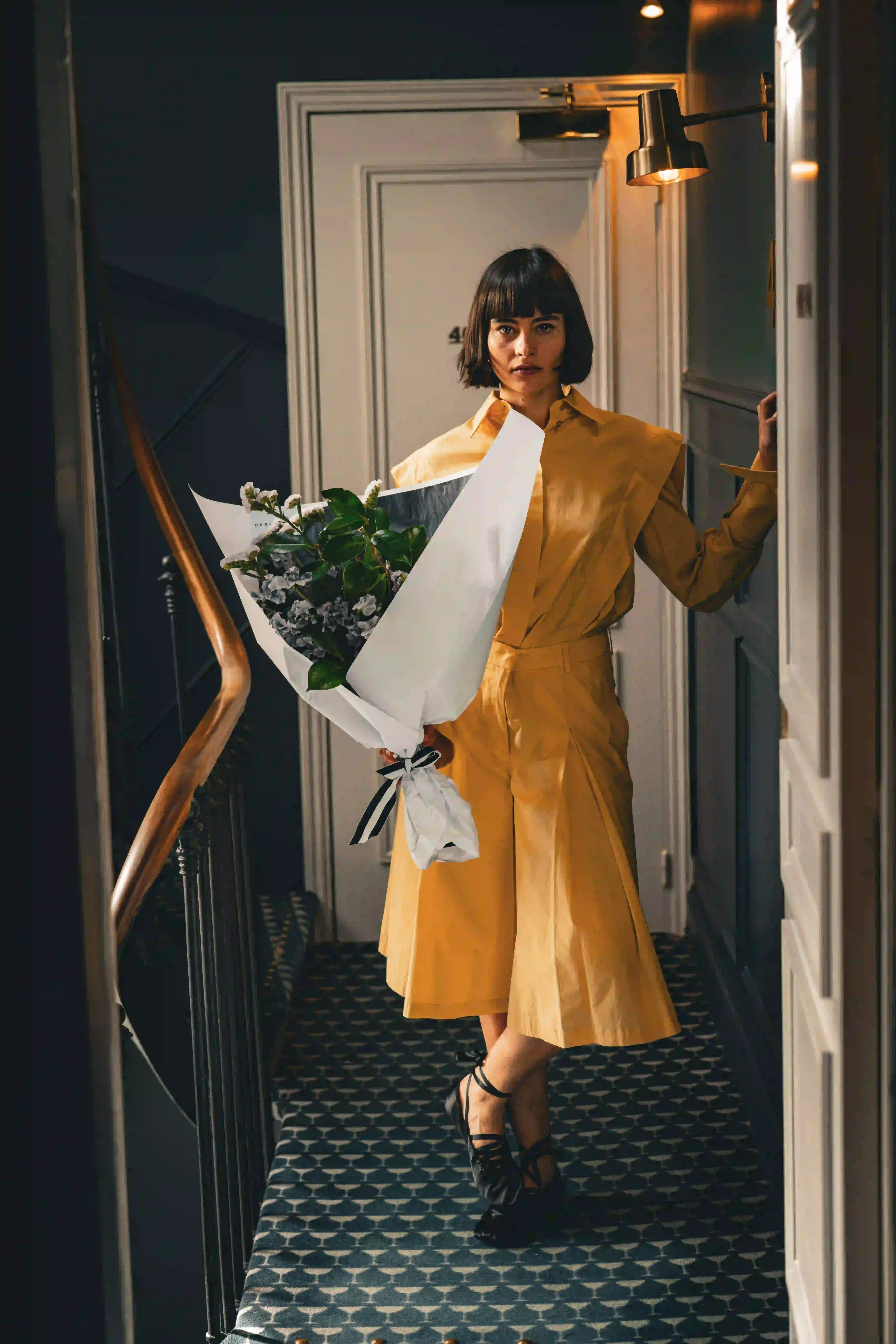 Woman in yellow outfit standing in hallway holding a large bouquet wrapped in white paper.