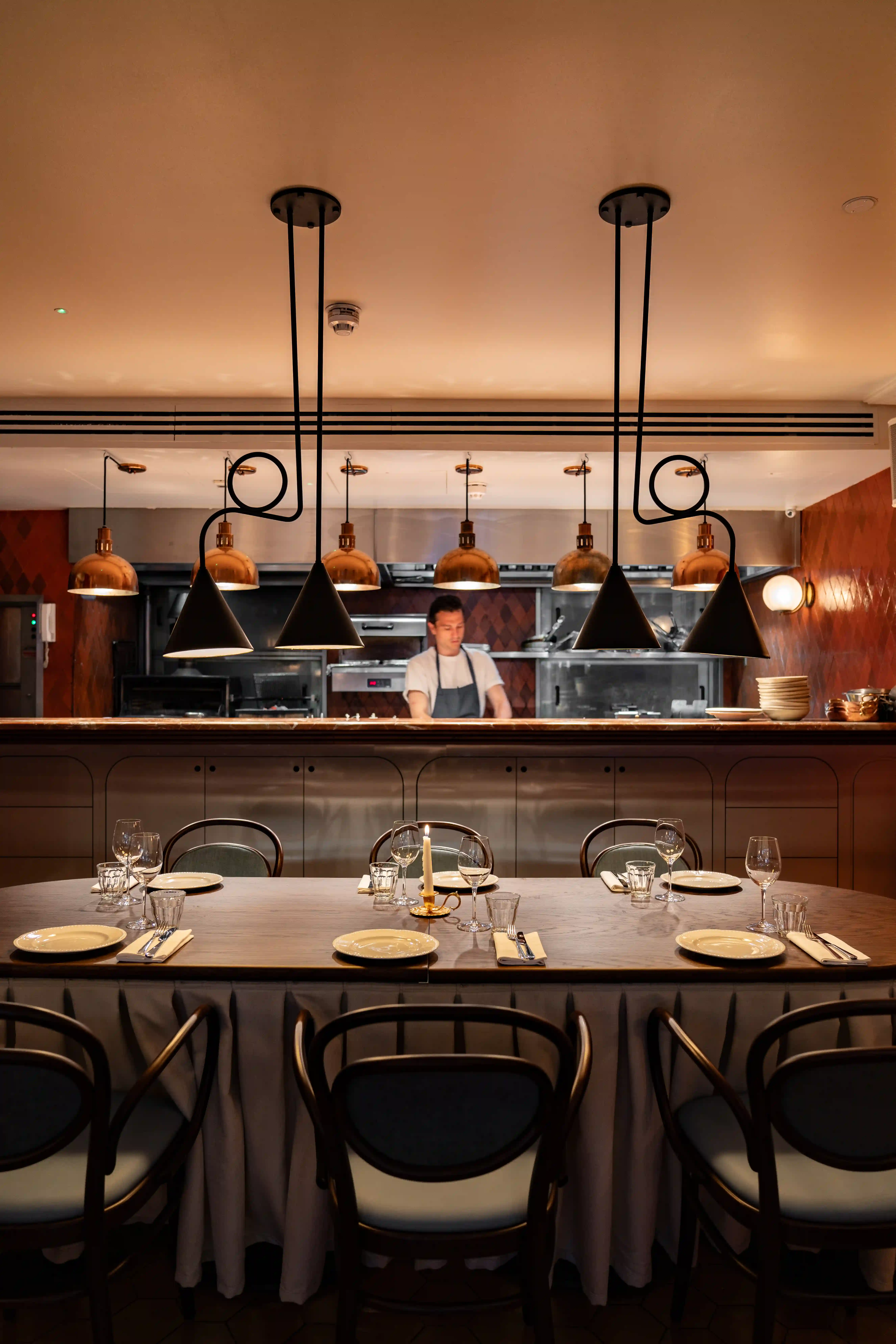 Table set for six with plates, glasses, and a candle in front of a chef in a warmly lit kitchen.