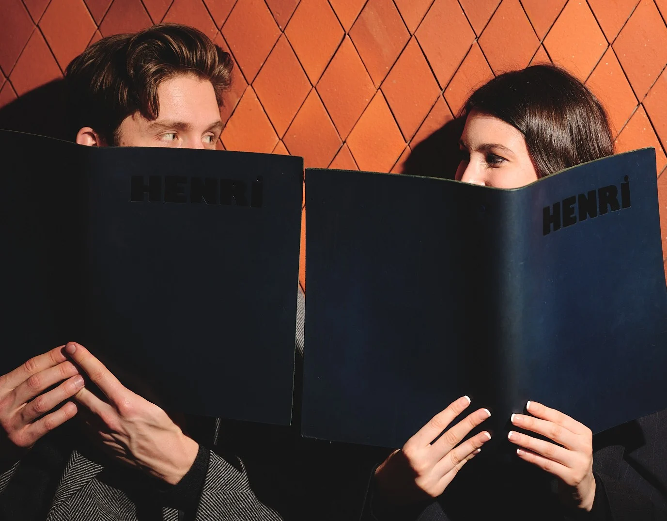 Man and woman hiding their faces behind menus with Henri printed, looking at each other.