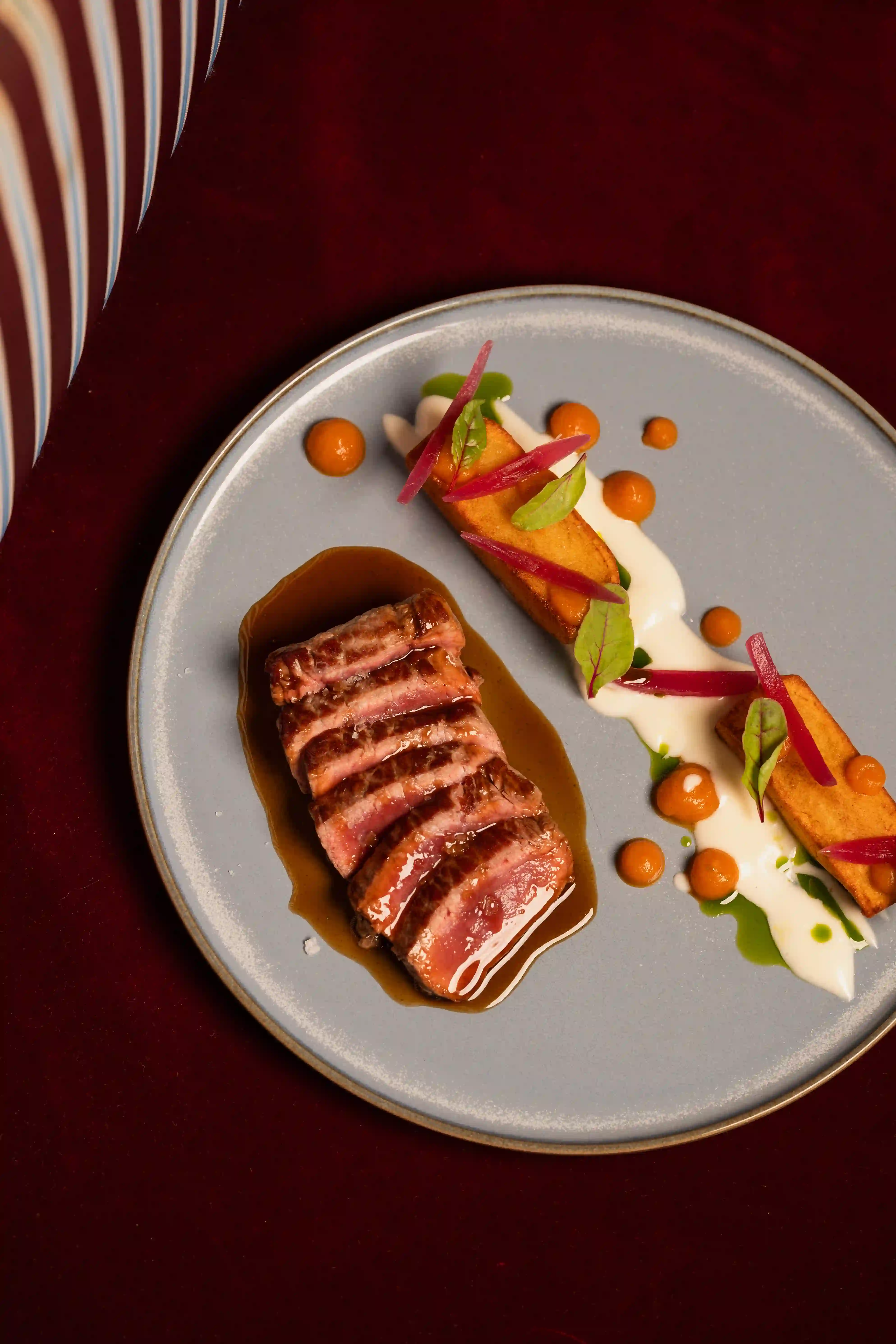 Slices of medium-rare steak with sauce, paired with rectangular fried pieces and garnishes on a gray plate.